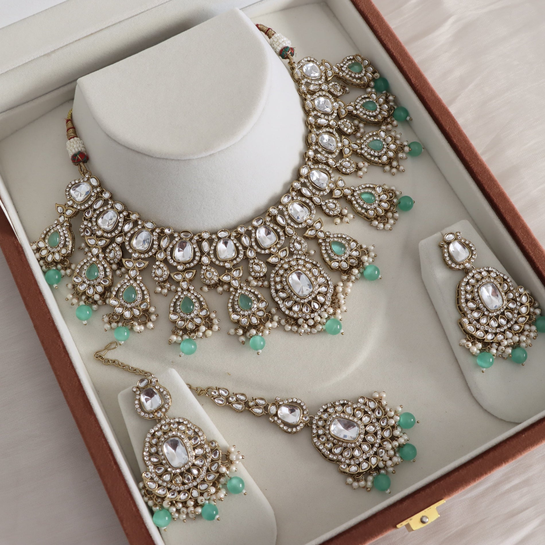 An ornate Indian-style jewelry set displayed inside a brown presentation box. The set includes a statement necklace on a white mannequin stand, matching drop earrings, and a matching bracelet, all featuring gold-plated metalwork, clear stones, and small green bead accents.