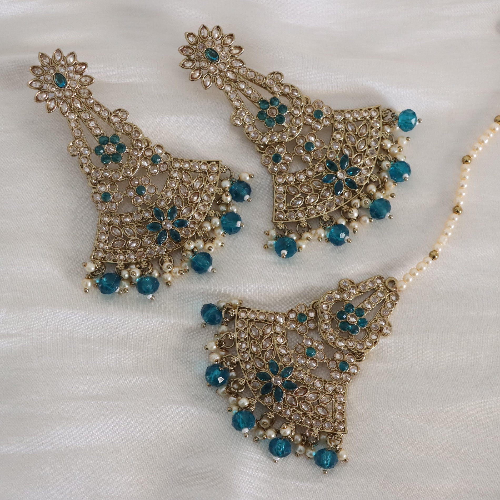 Gold-toned traditional Indian jewelry set featuring a large pendant, matching earrings, and a pearl-bead chain, embellished with turquoise blue stones and small white pearls, displayed on a white textured fabric background.