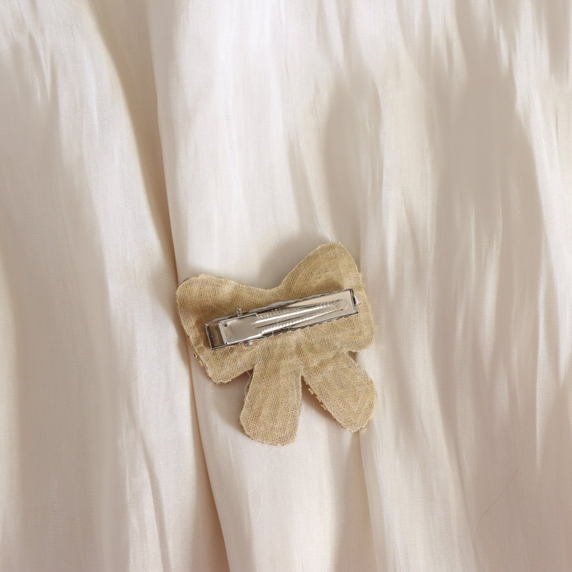 A close-up of a beige fabric bow hair clip with a silver metal spring clip, pinned onto a cream-colored textured fabric.
