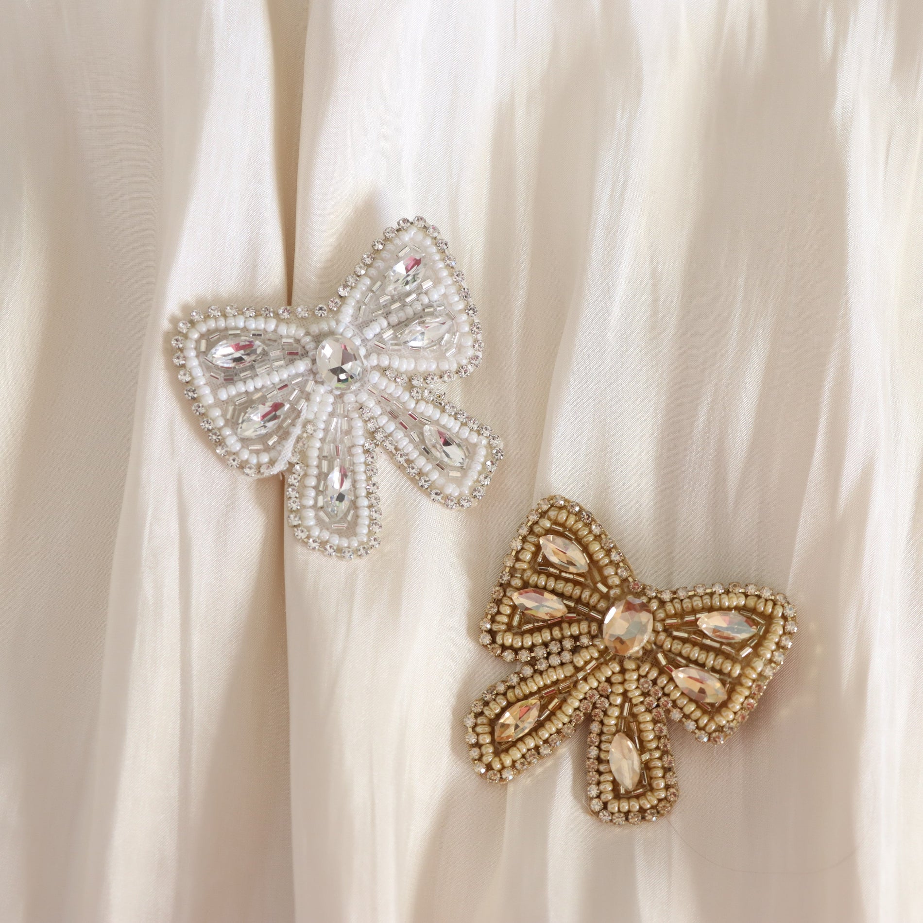 Two decorative bow-shaped brooches with beaded detailing and rhinestones, one silver-toned and one gold-toned, displayed on a cream-colored draped fabric background.