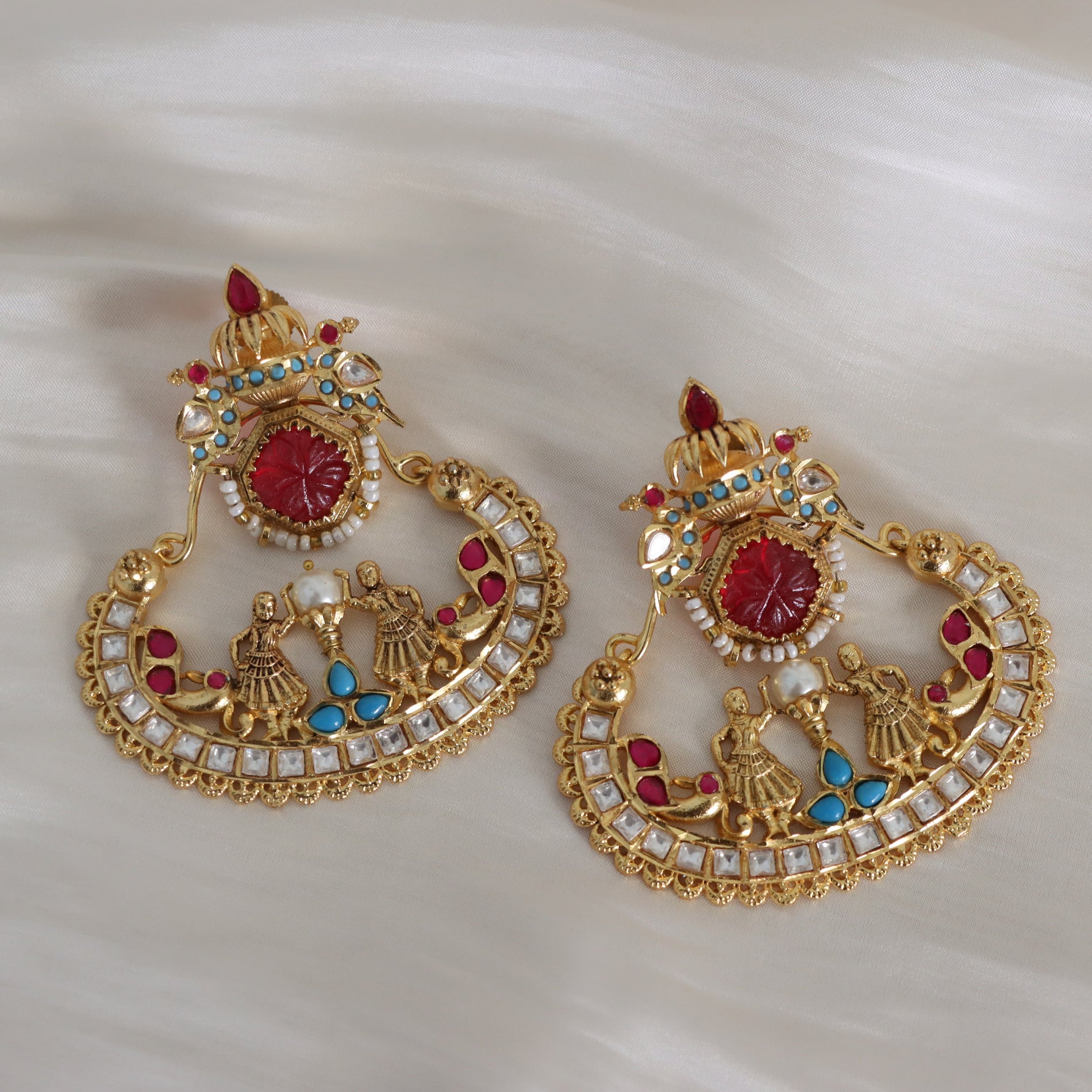 A pair of ornate gold-toned Indian chandbali earrings decorated with red cabochon stones, white faux pearls, and small blue and red accent stones, displayed on a soft white fabric background.