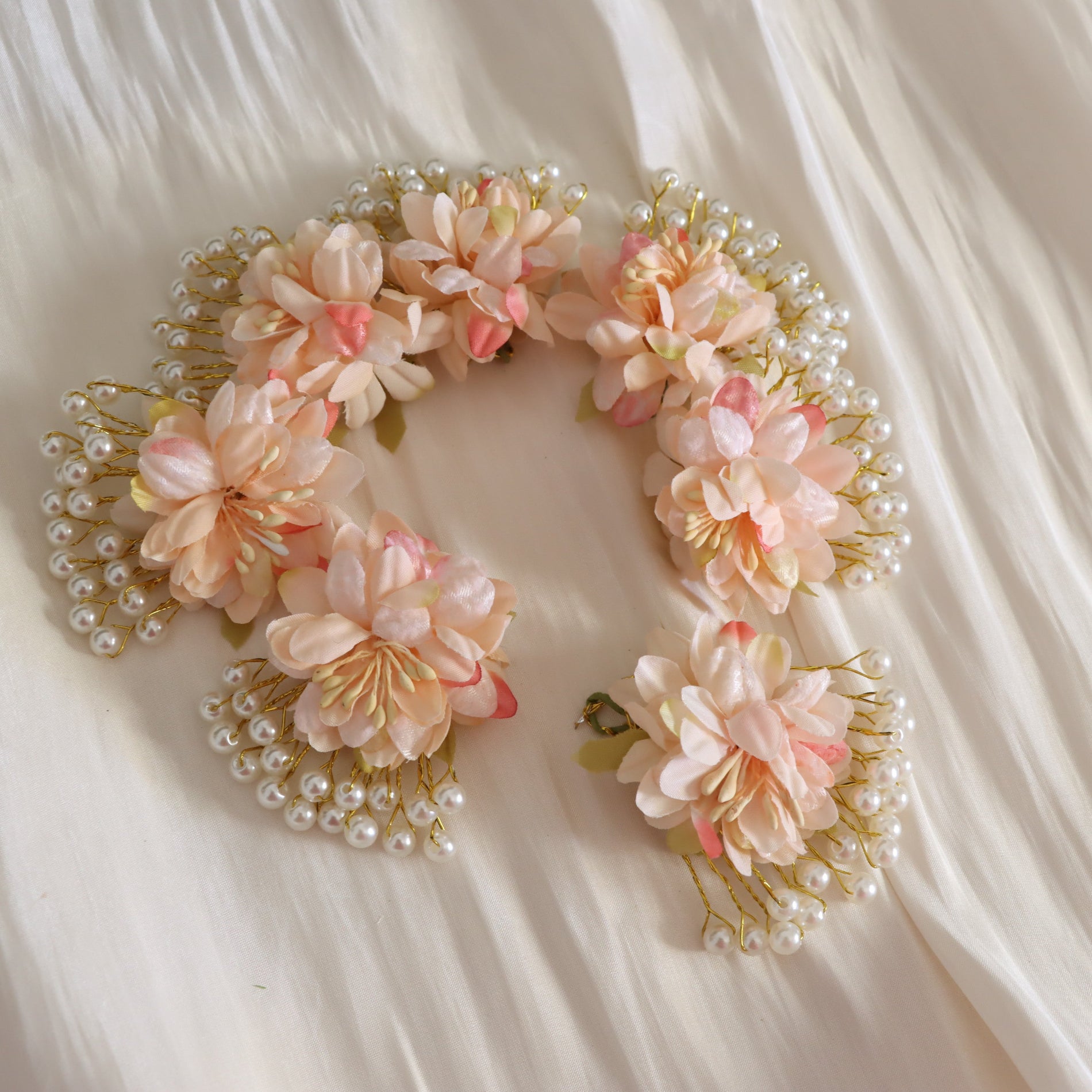 A decorative floral headpiece with layered peach and cream faux flowers, pearl-like beads, and gold-toned wire accents, arranged in a circular shape on a soft cream fabric background.