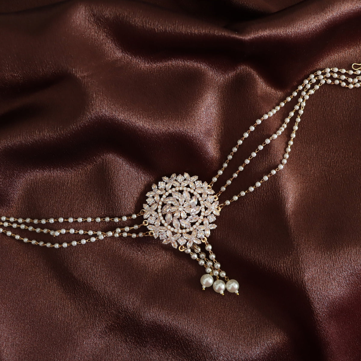 A double-strand pearl choker necklace with a large, ornate round pavé centerpiece and dangling pearl beads, displayed on a draped dark brown satin fabric.