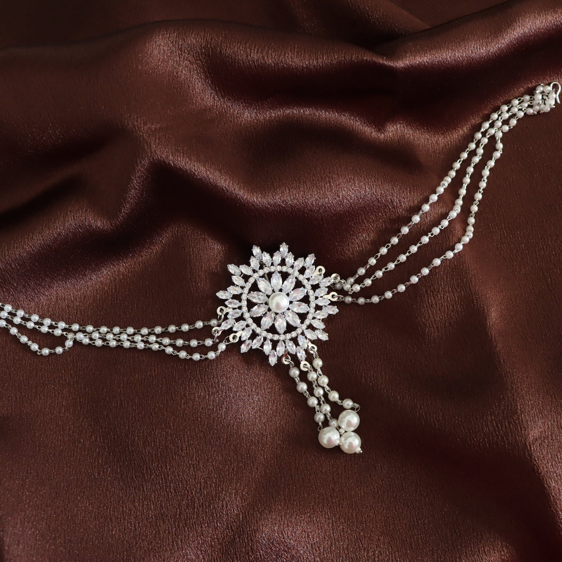 A silver-toned double-strand chain necklace with a central floral brooch set with clear crystals and dangling pearl-like beads, displayed on a draped dark brown satin fabric.