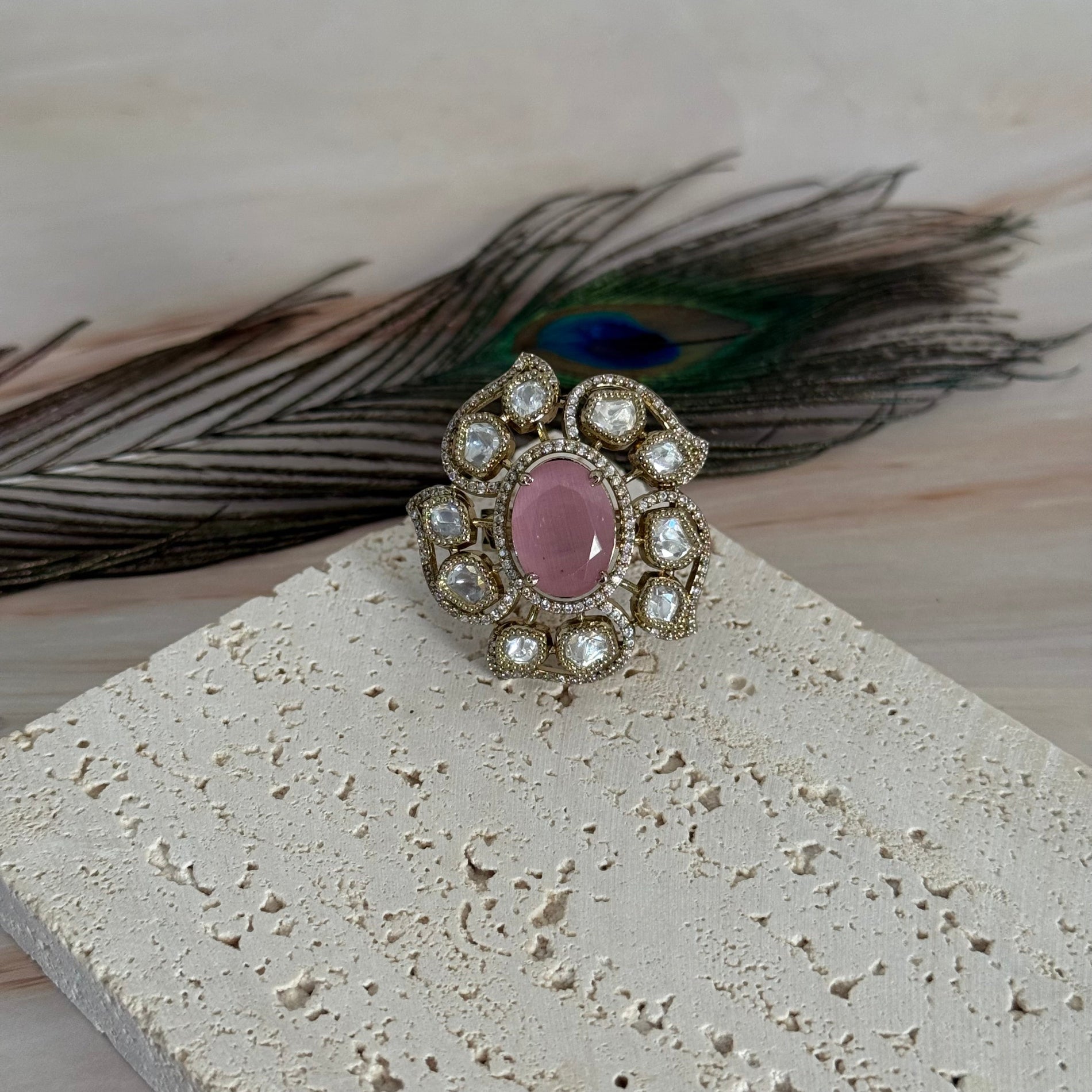 Decorative ring with a pink gemstone and pearl-like stones on a textured surface with peacock feathers.