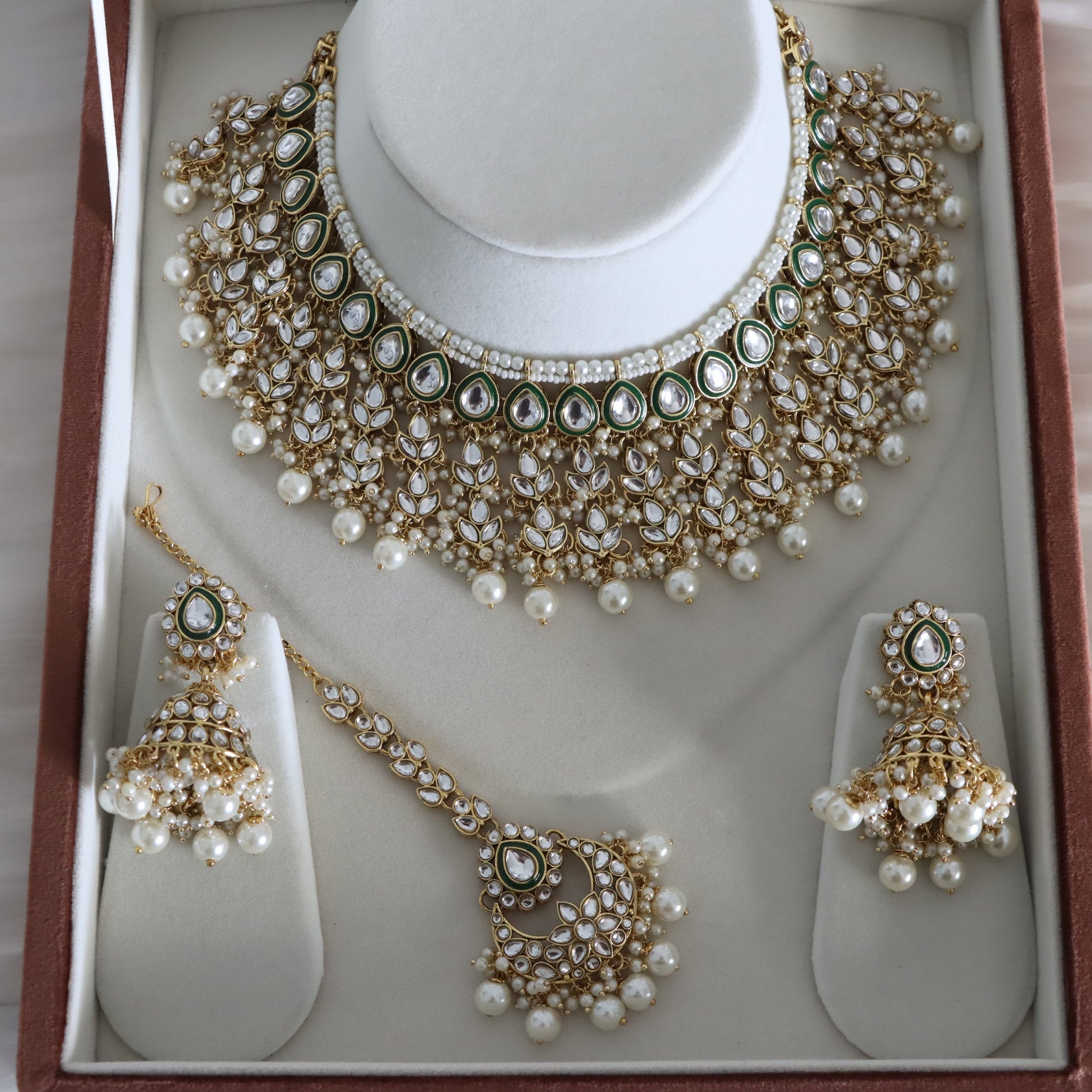 Gold and white pearl jewelry set displayed on a white jewelry stand inside a brown presentation box, including a choker necklace, matching earrings, and a maang tikka.