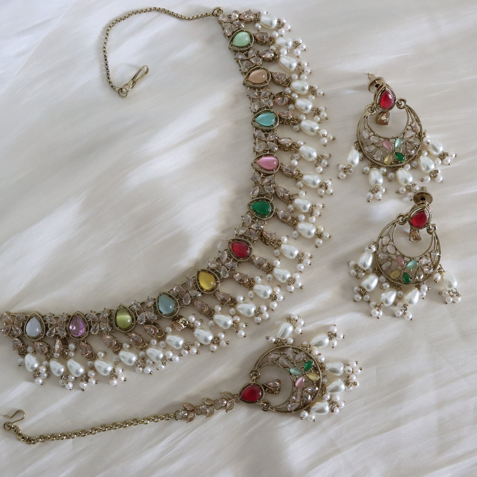 Gold-plated Indian jewelry set featuring a necklace, earrings, and a maang tikka, embellished with white faux pearls and colorful gemstones in red, green, blue, pink, and yellow, displayed on a white fabric background.