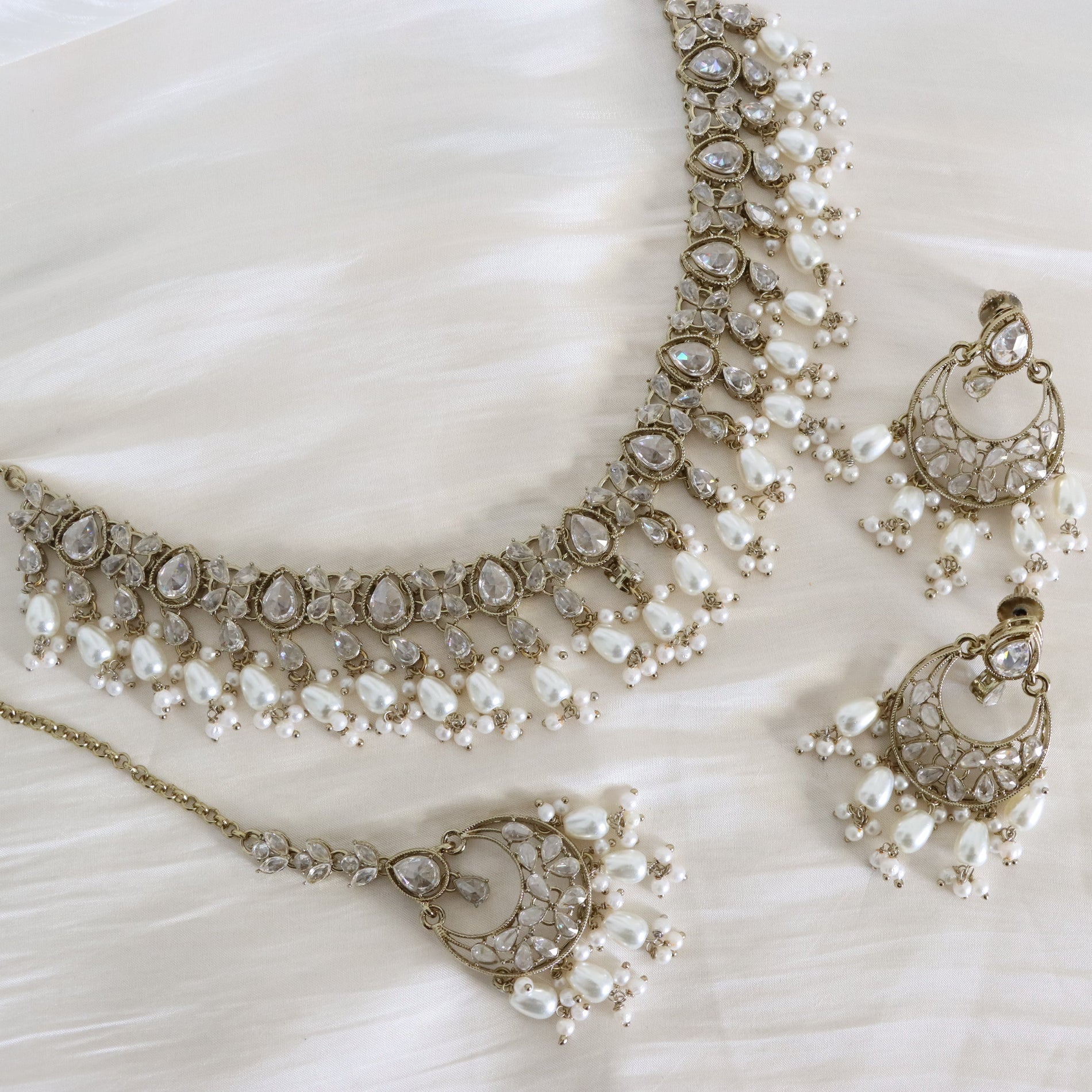 An ornate jewelry set laid out on a soft white fabric background, featuring a necklace, matching earrings, and a pendant in gold-tone metal with clear stones and white pearl-like drops.