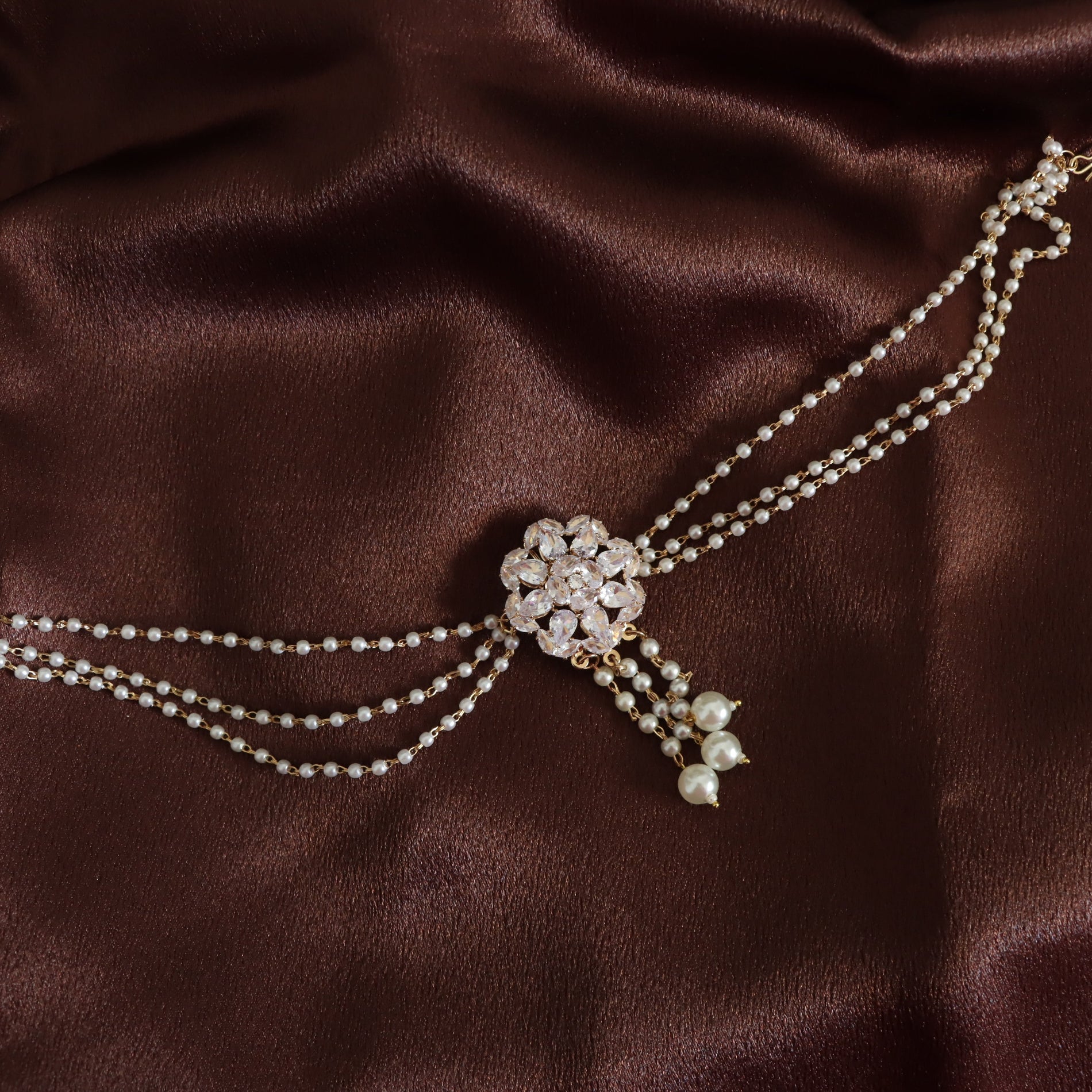 A double-strand pearl choker necklace with a central rhinestone flower cluster and dangling pearl beads, displayed on a draped dark brown satin fabric background.