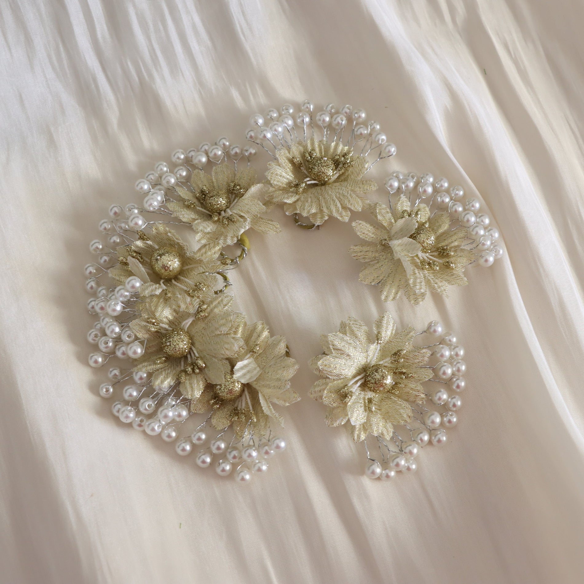 A set of three matching hair clips decorated with cream-colored fabric flowers, gold glitter centers, and rows of small white faux pearls, arranged on a draped cream satin fabric background.