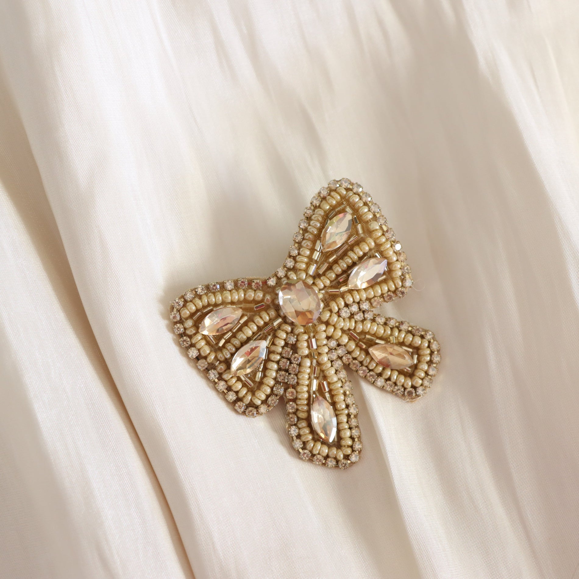 A bow-shaped beaded brooch with iridescent pale pink rhinestones, displayed on a draped cream-colored fabric background.