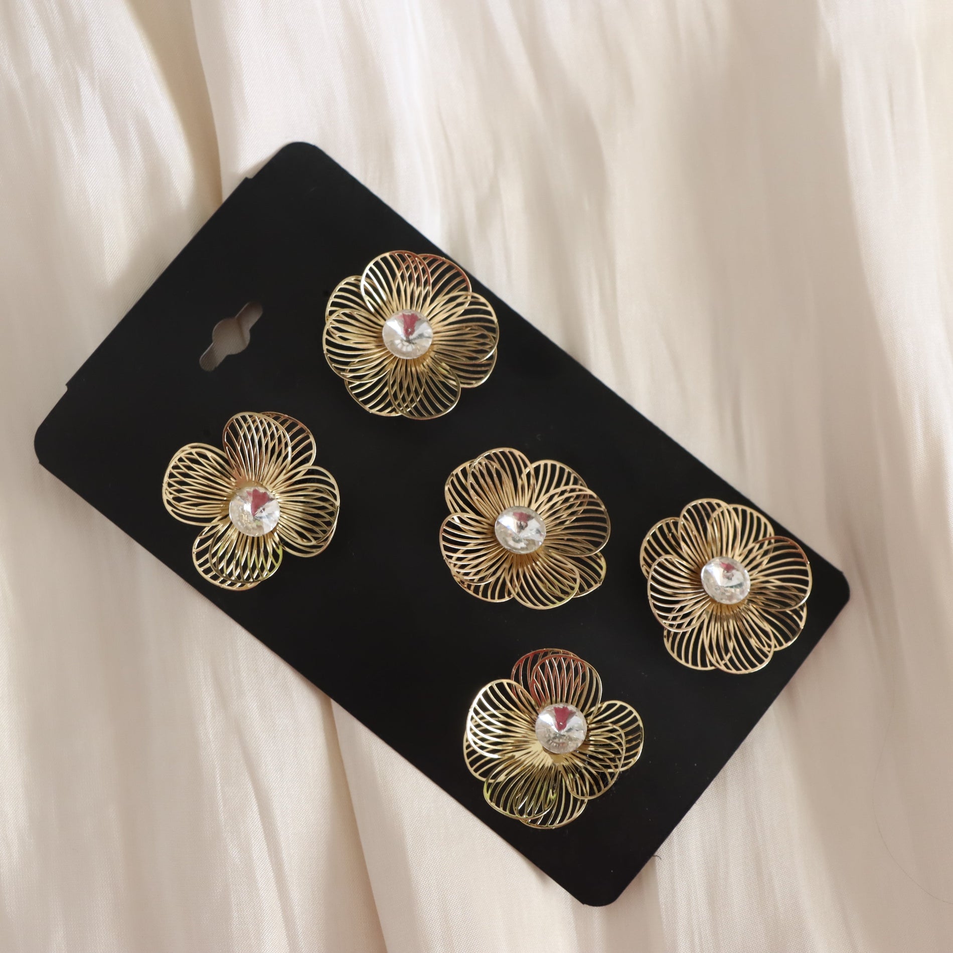 Five gold-tone flower-shaped hair clips with clear rhinestone centers, displayed on a black backing card over a cream-colored fabric background.