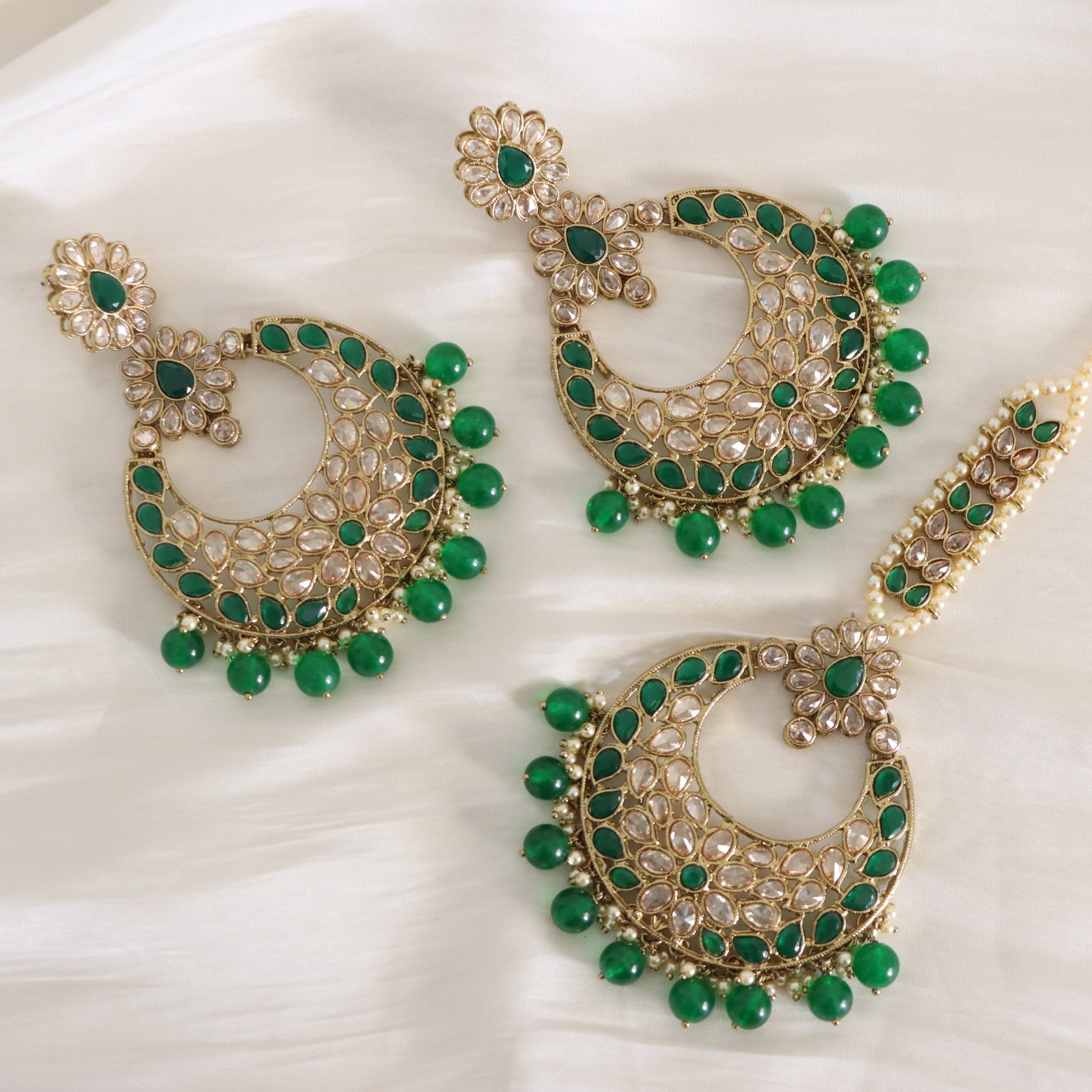 Gold-plated crescent-shaped Indian earrings with green stone accents and clear rhinestone detailing, arranged on a white fabric background.
