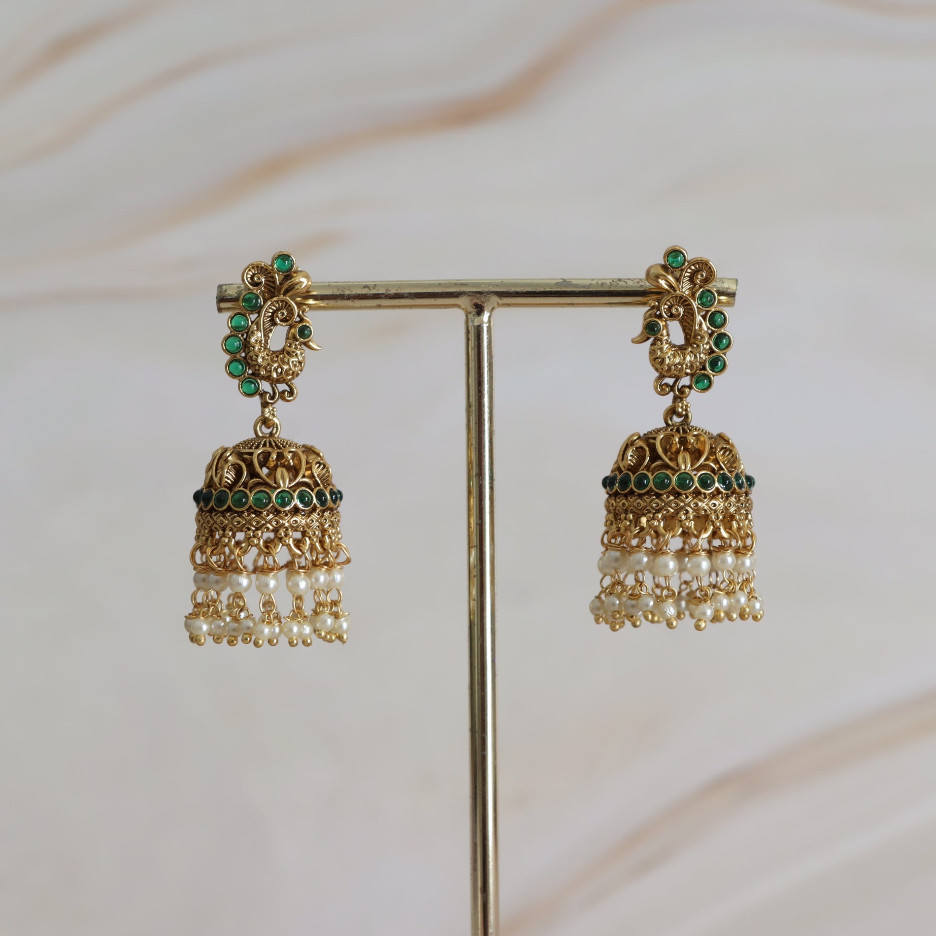 Gold earrings with pearl and green gemstone details on a stand against a light background