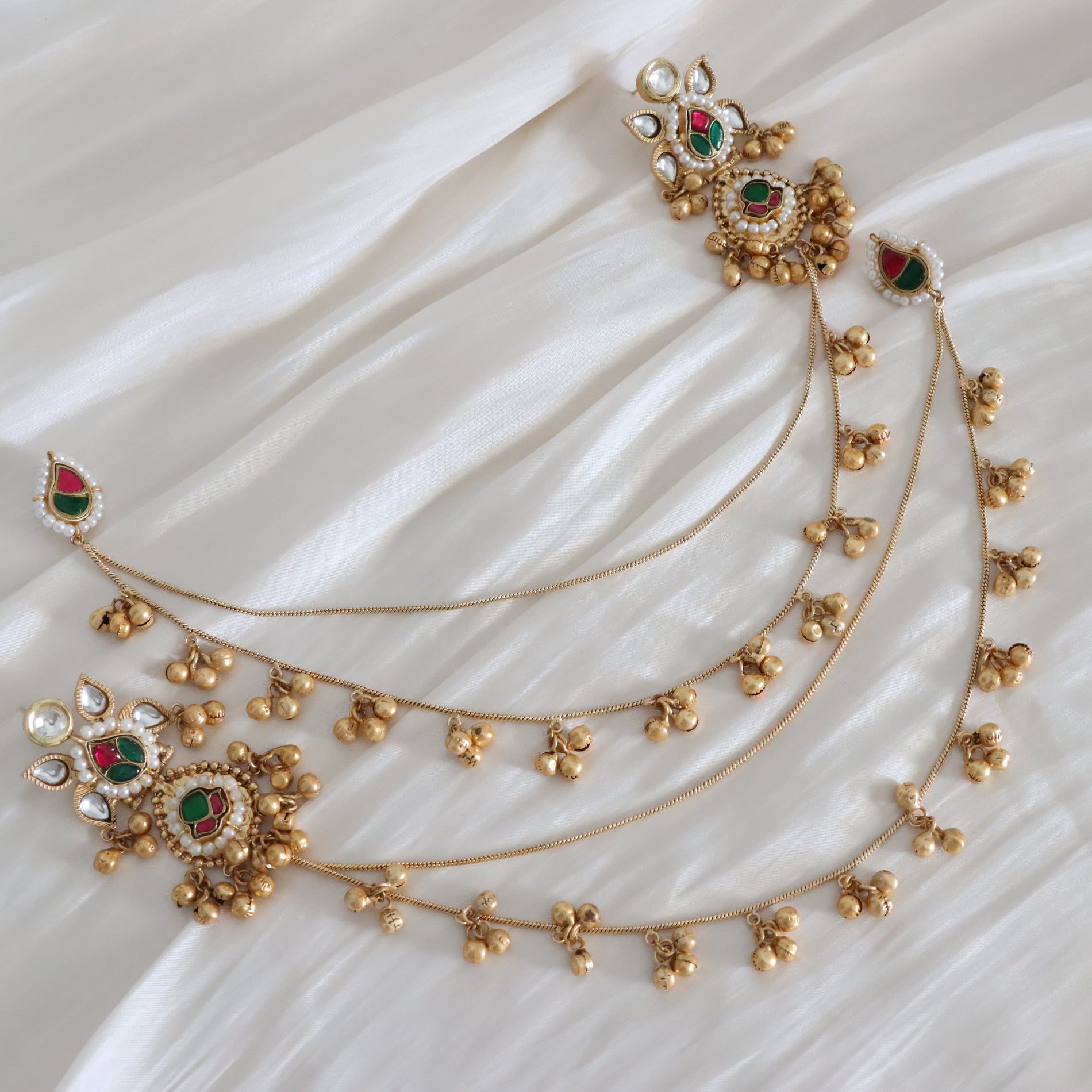 Gold-toned traditional jewelry set with intricate filigree, small bell charms, and red and green stone accents, arranged on a draped white fabric background.