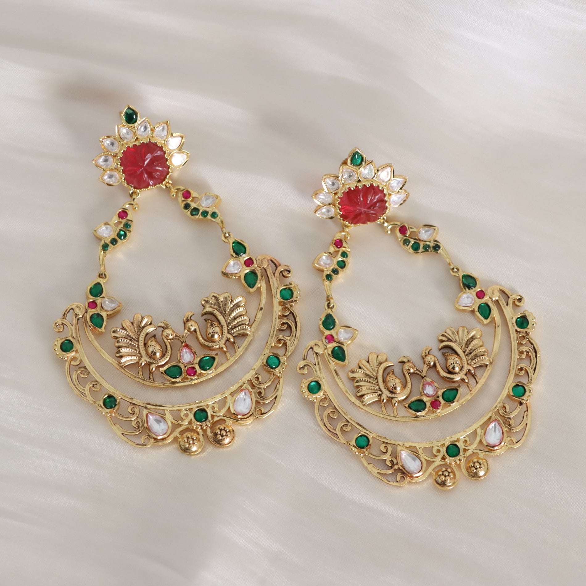 A pair of ornate gold-toned traditional Indian earrings decorated with red stones, green stones, and white stones, displayed on a soft white fabric background.