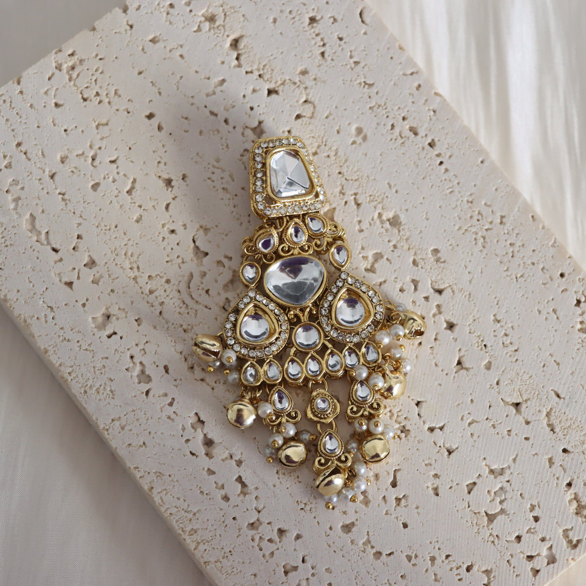 An ornate gold-toned chandelier earring with clear stones and small pearl-like accents, displayed on a textured beige stone slab.