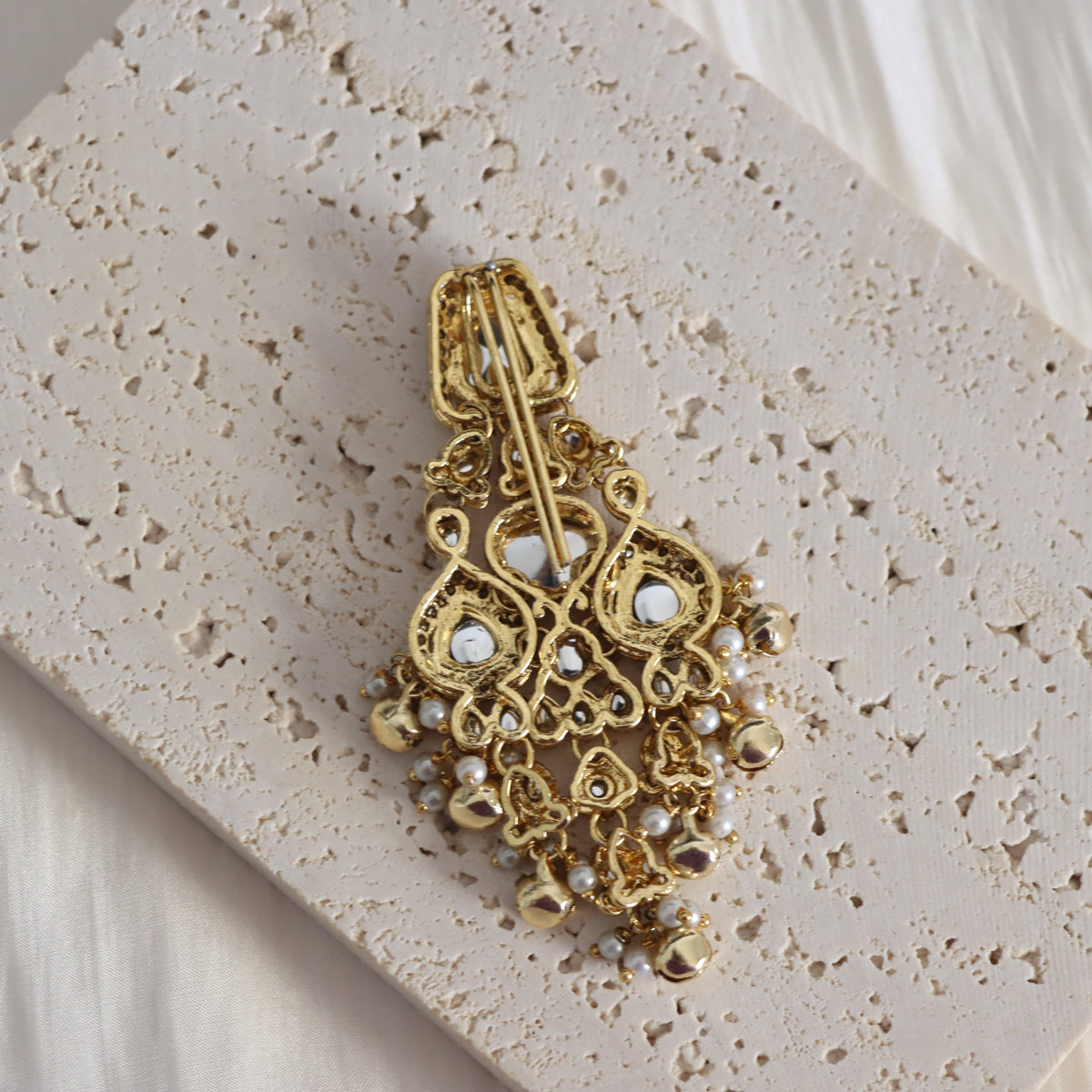 An ornate gold-toned statement earring with intricate filigree detailing and small white pearl-like accents, displayed on a textured beige stone tile.