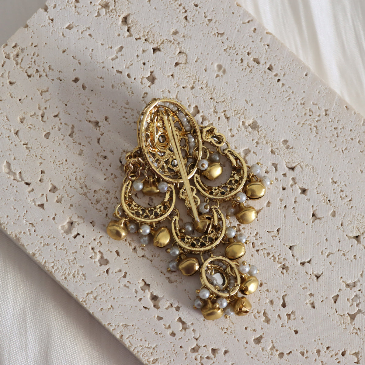 An ornate gold-toned vintage brooch with filigree detailing, small pearl-like beads, and dangling gold bells, displayed on a textured beige stone tile.