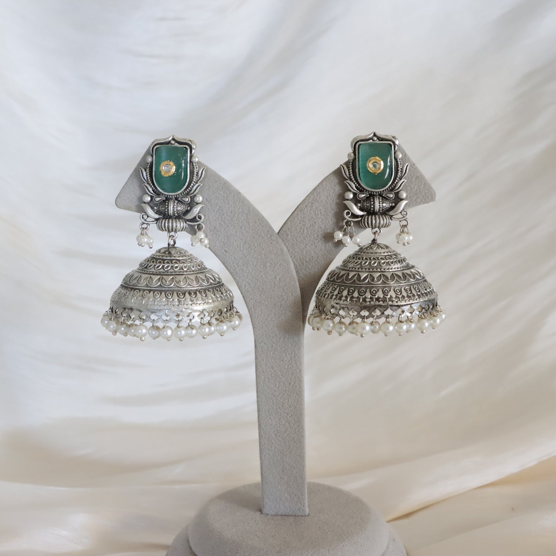 A pair of traditional Indian silver jhumka earrings with green stone and gold foil accents, displayed on a gray velvet stand against a draped white fabric background.
