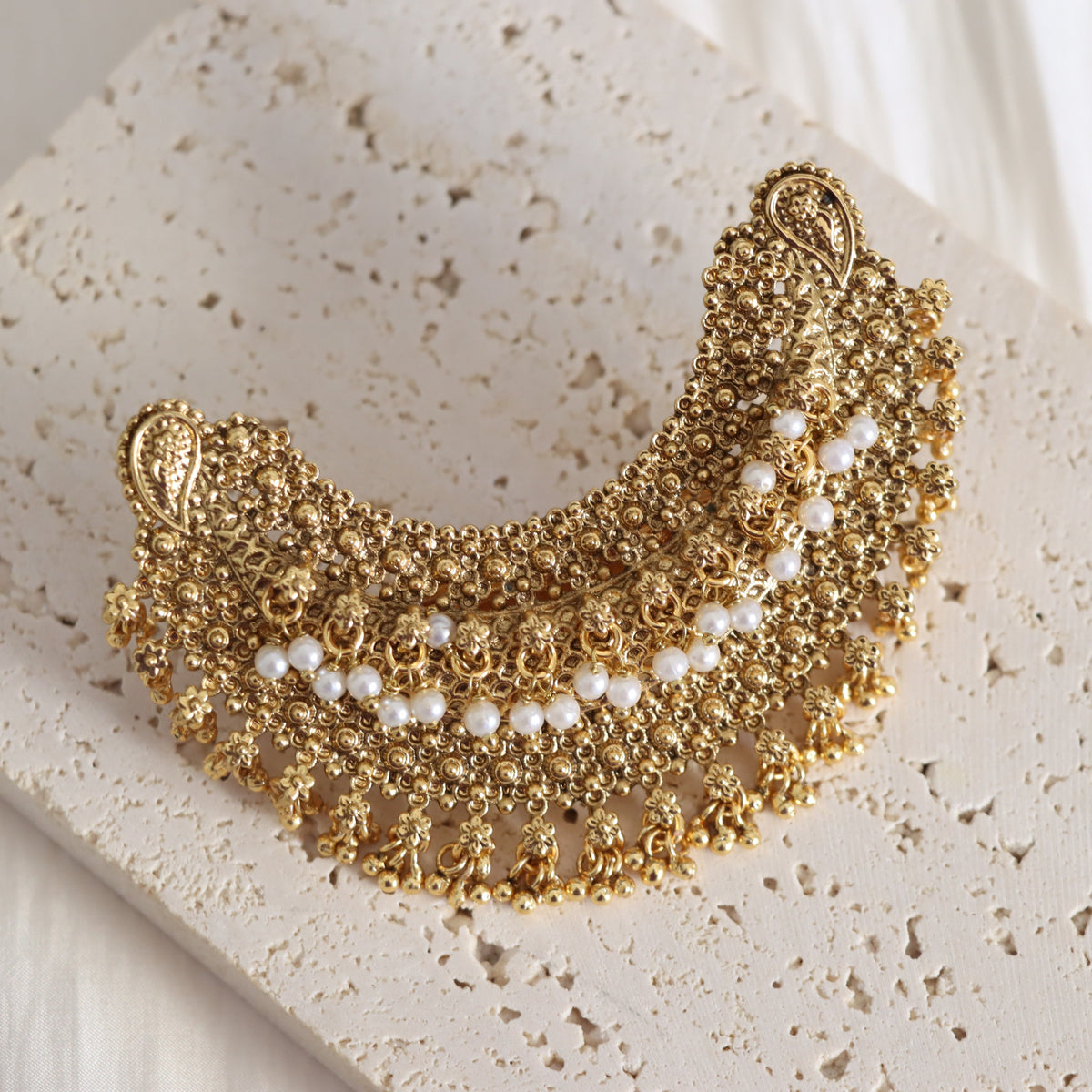 An ornate gold crescent-shaped collar brooch decorated with intricate floral filigree, small pearls, and dangling floral details, displayed on a textured beige stone slab.