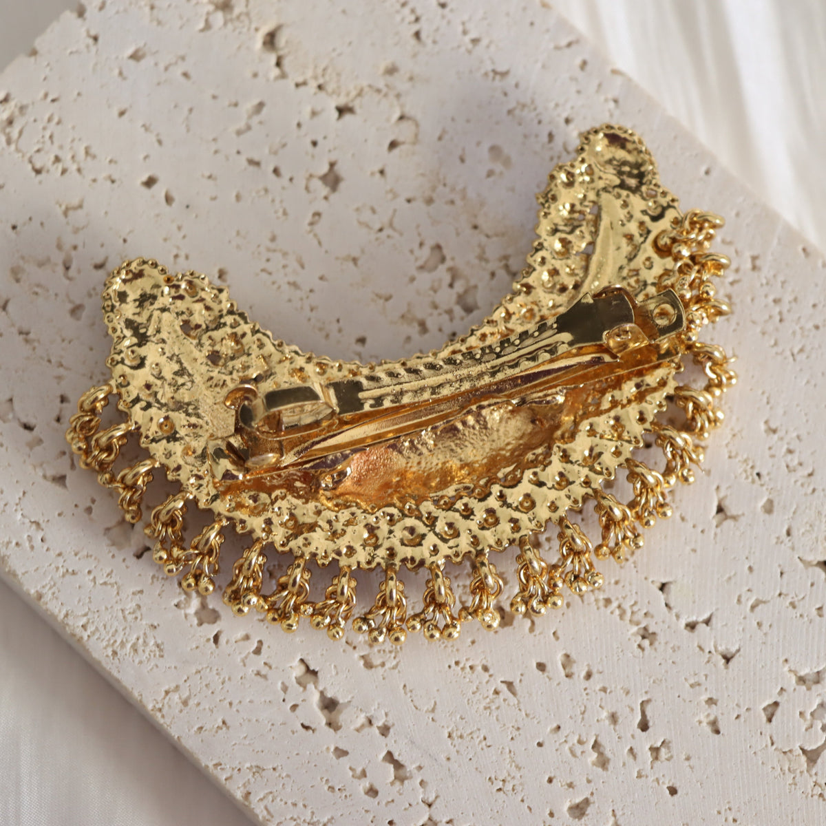 A gold-tone metal hair claw clip with an ornate filigree lace design and dangling chain fringe, displayed on a textured beige stone tile.