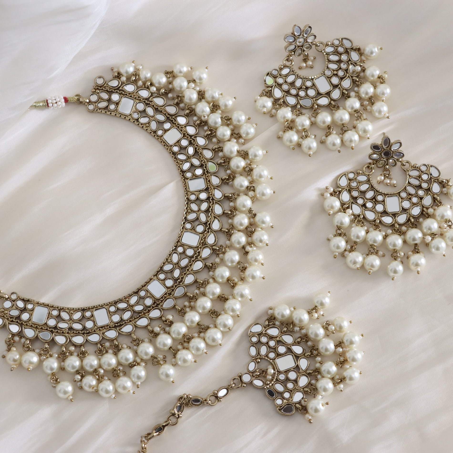 An ornate Indian ethnic jewelry set featuring a crescent-shaped necklace, matching earrings, and a maang tikka, all embellished with white faux pearls and silver-toned metalwork, displayed on a draped white fabric background.