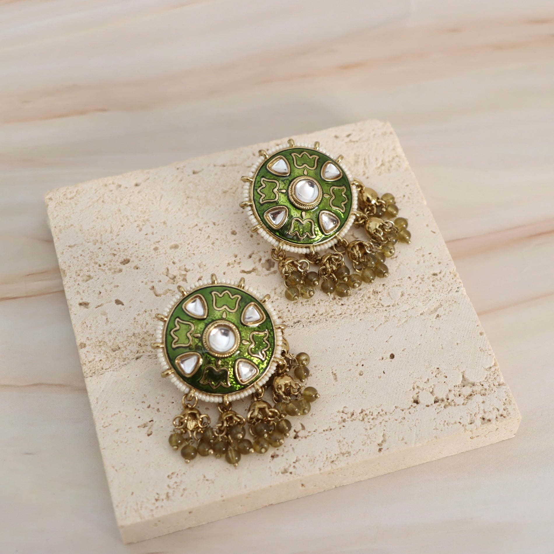 A pair of green and gold Indian-style statement earrings with white stone accents and dangling bead chains, displayed on a textured beige stone tile over a light wood background.