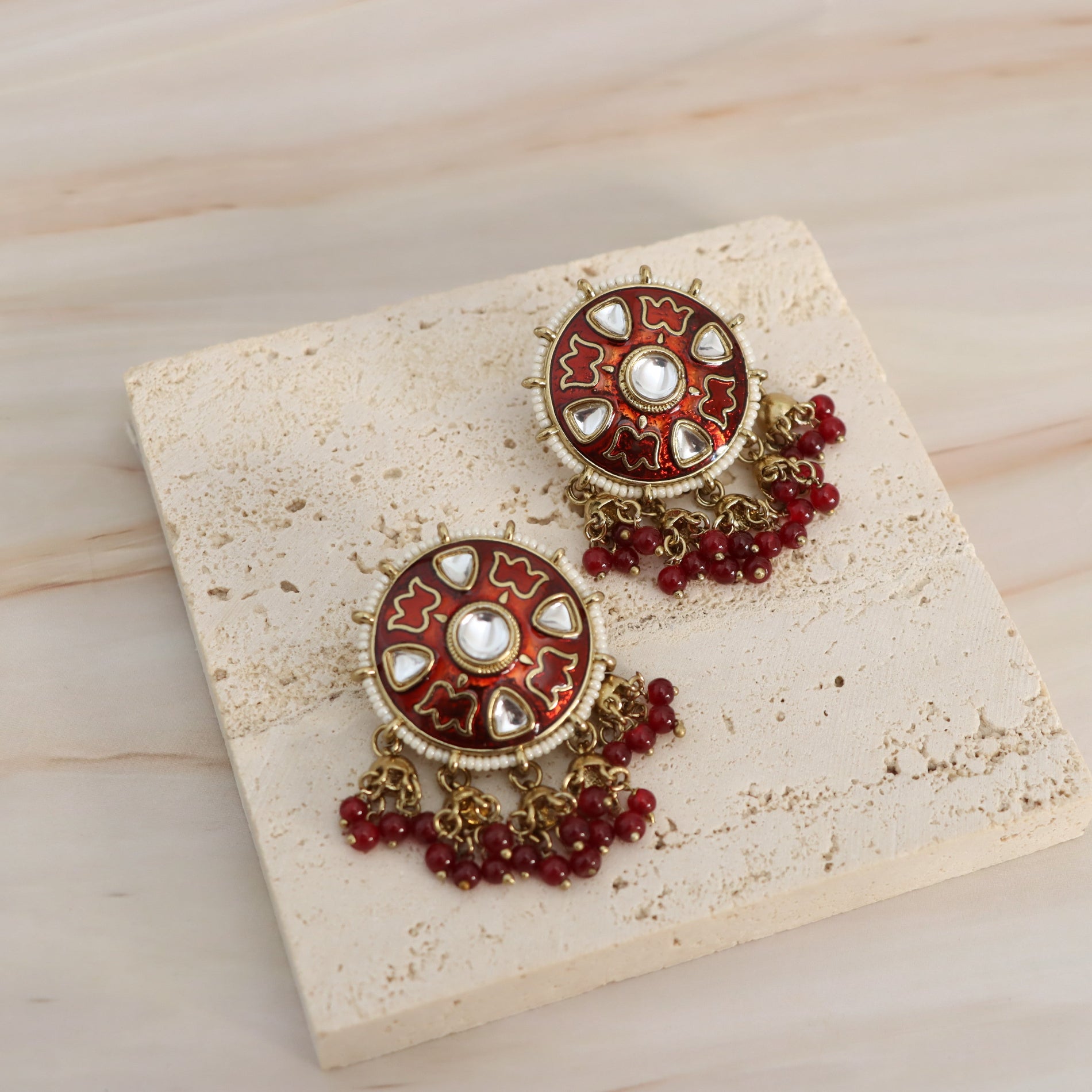A pair of ornate Indian-style chandelier earrings displayed on a textured cream stone tile over a light wood background. The earrings feature circular red and gold enamel studs with silver-toned metal details and dangling red bead chains.