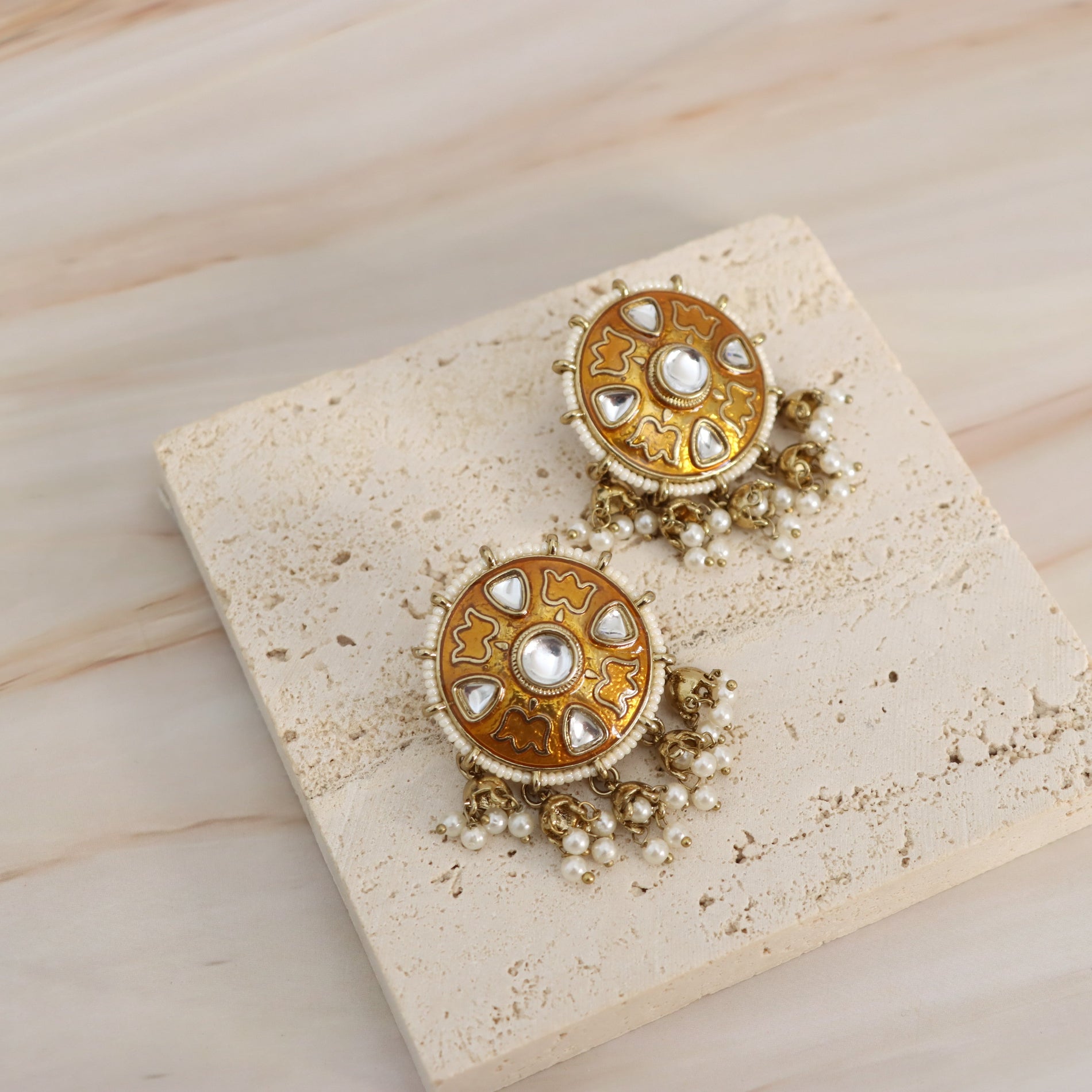 A pair of ornate gold-toned statement earrings with white pearl-like accents and dangling bead chains, displayed on a textured beige stone slab over a light wood surface.