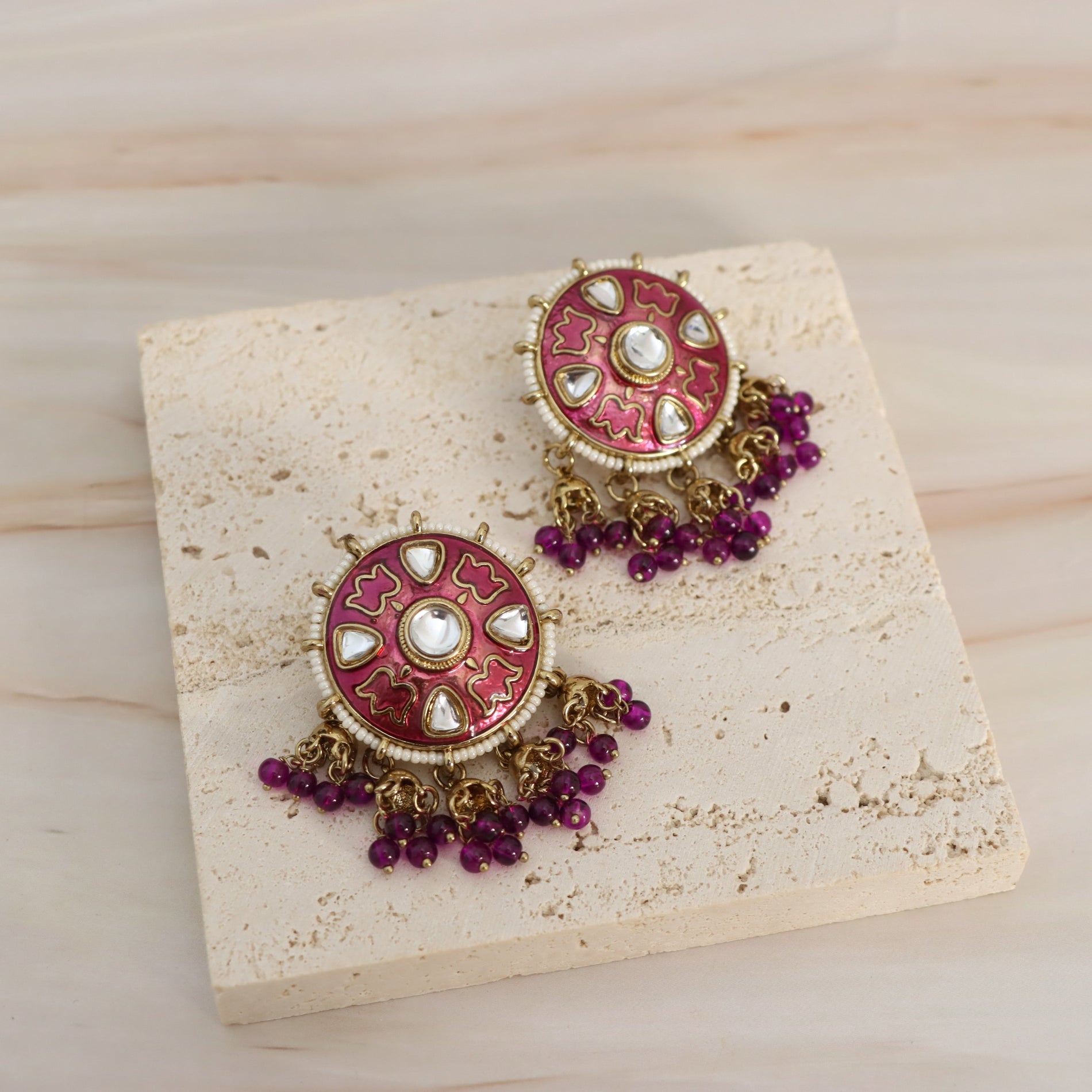 A pair of ornate Indian-style earrings with circular pink and gold-plated studs, decorated with white stones and dangling clusters of purple beads, displayed on a textured beige stone slab over a light wood background.