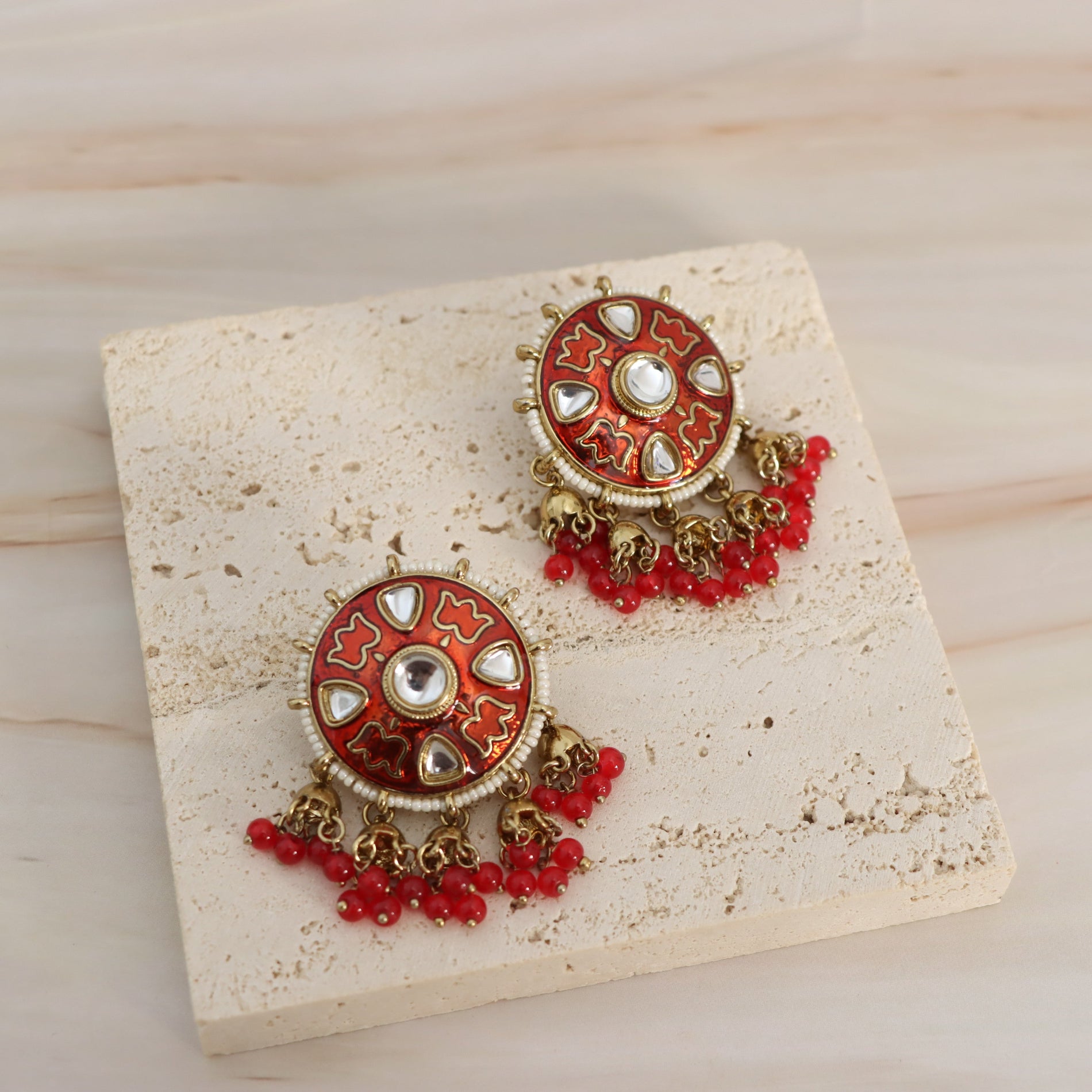 A pair of ornate Indian-style dangle earrings displayed on a textured beige stone slab over a light wood background. The earrings feature circular red and gold enamel tops with white stone accents, hanging gold-toned chains, and clusters of small red beads.