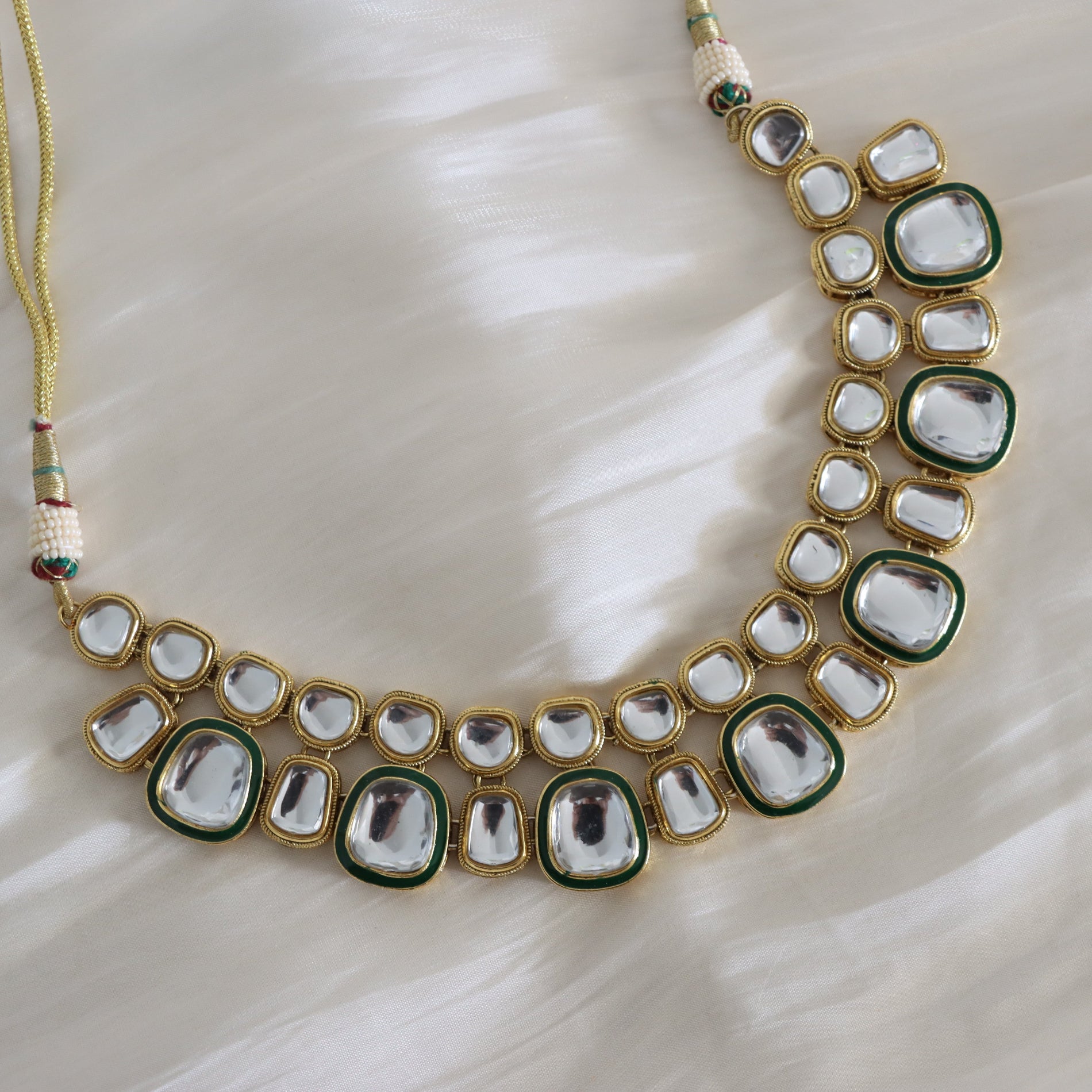 A gold-plated statement necklace featuring large clear glass cabochons bordered by dark green enamel and smaller clear stones, displayed on a soft white fabric background.