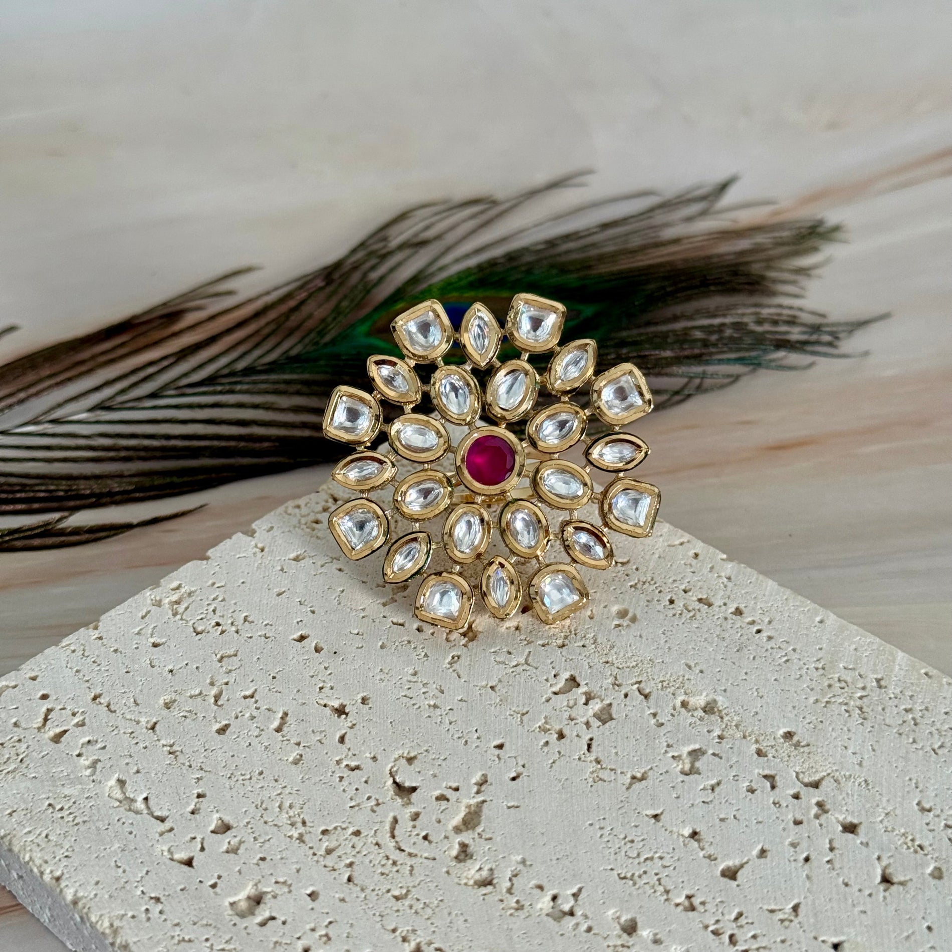 Decorative ring with gemstones and a red stone on a textured surface with peacock feathers.