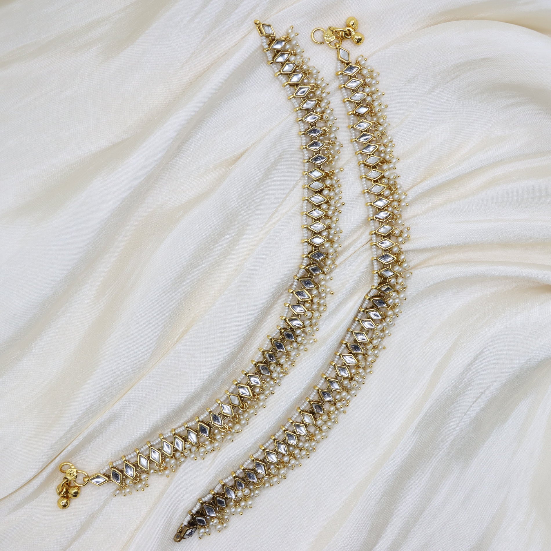 Gold-toned beaded ankle bracelets with clear stones, arranged on a draped cream fabric background.