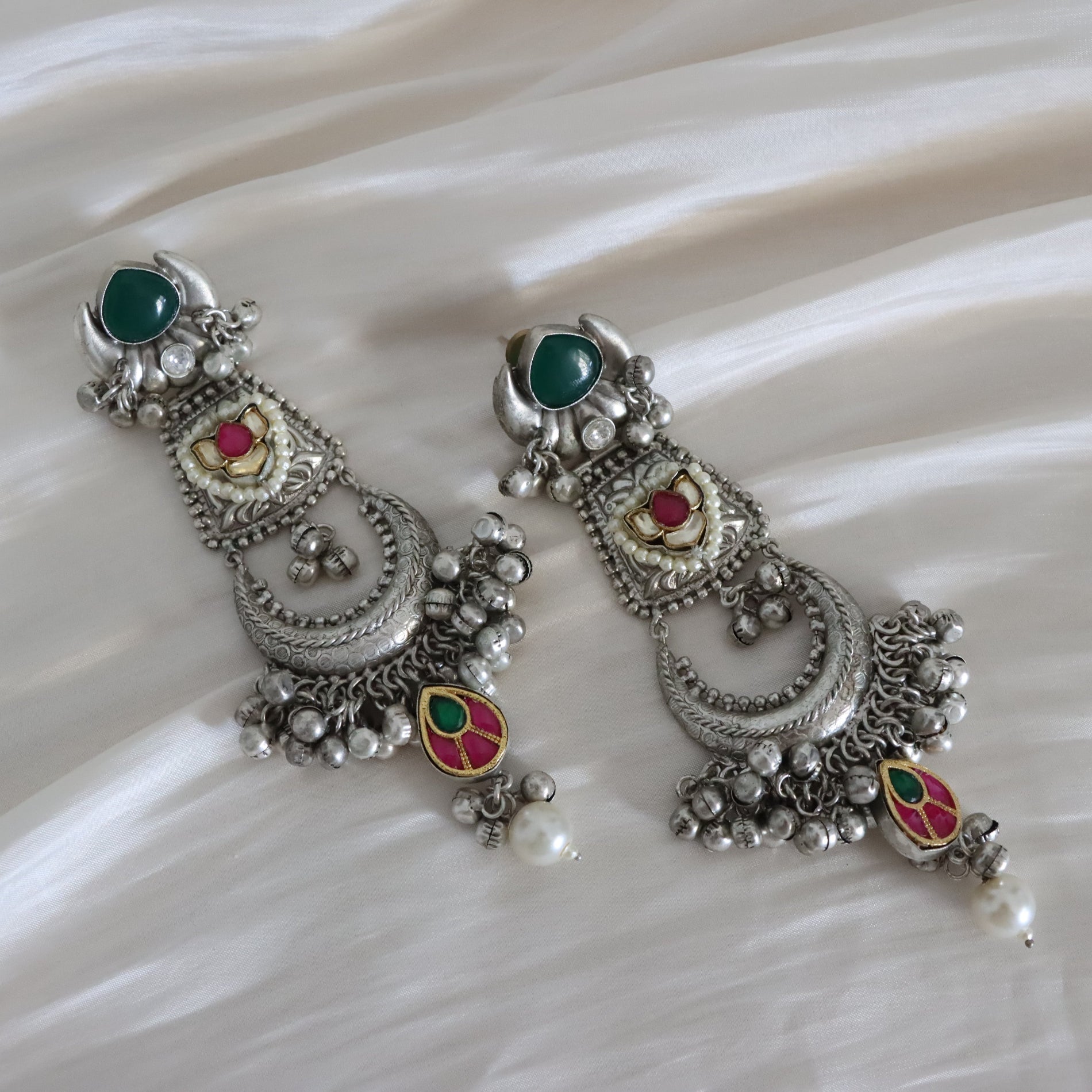 A pair of ornate silver-toned traditional Indian earrings with green stones, red and yellow enamel details, and white pearl-like drops, displayed on a draped cream satin fabric.
