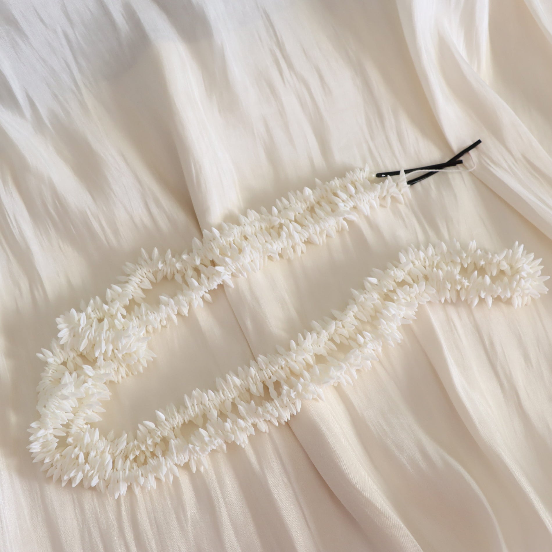 A white floral lei or garland laid out on a cream-colored fabric background, with two black bobby pins holding it in place.
