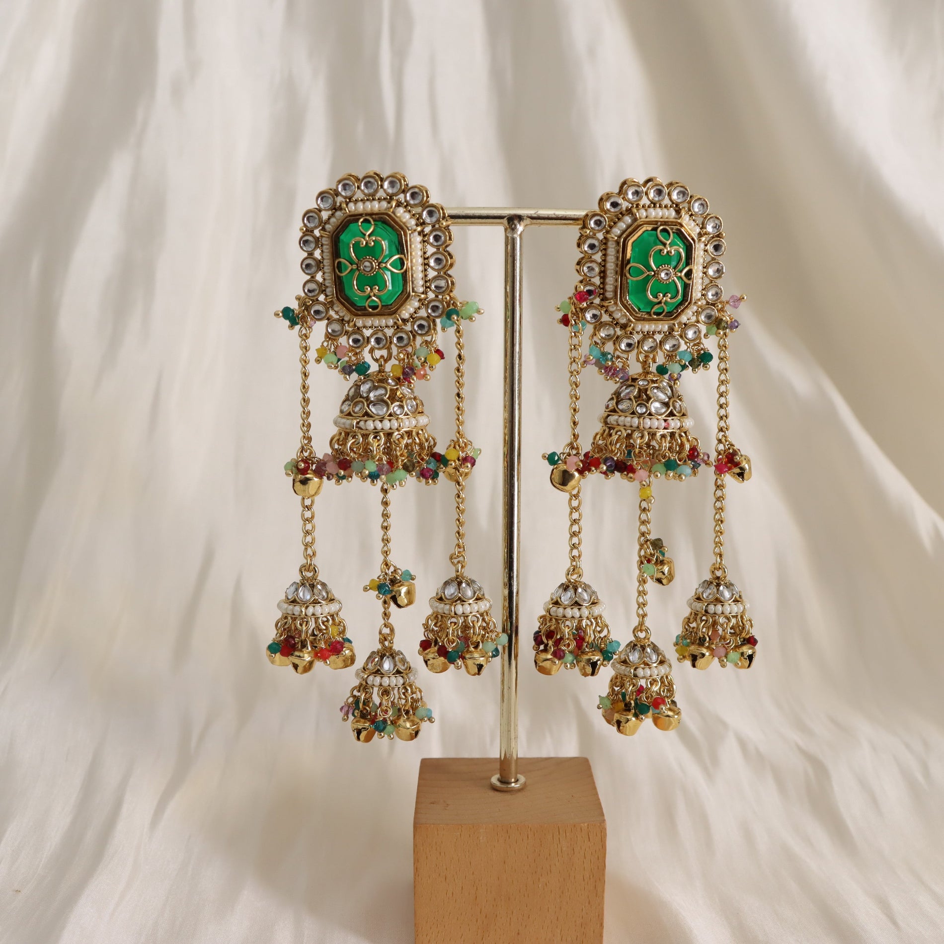 Ornate earrings with green gemstones and colorful beads on a stand against a white background