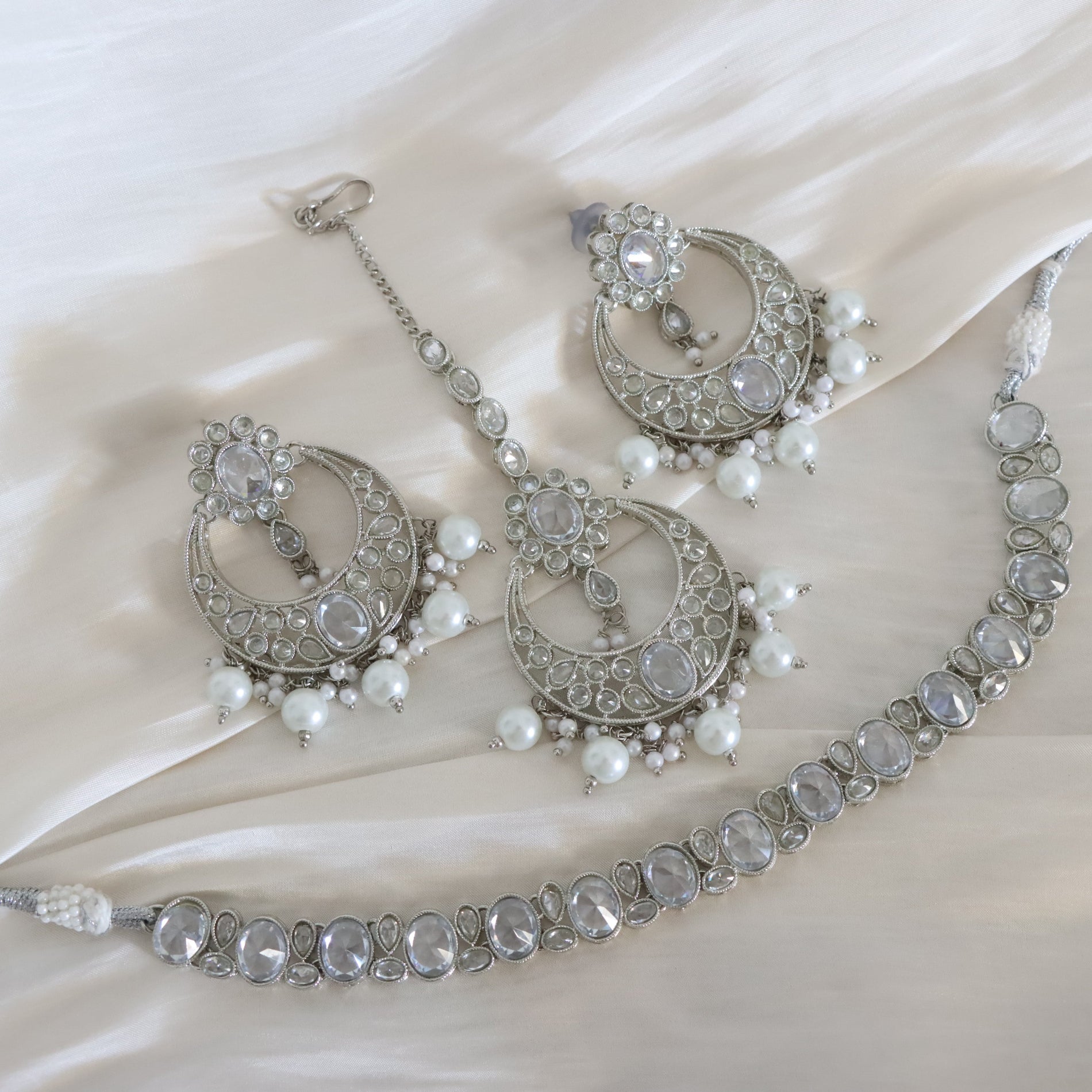 A silver-toned traditional jewelry set including a necklace, earrings, and a matching maang tikka, decorated with clear crystals and white pearls, arranged on a draped cream satin fabric.