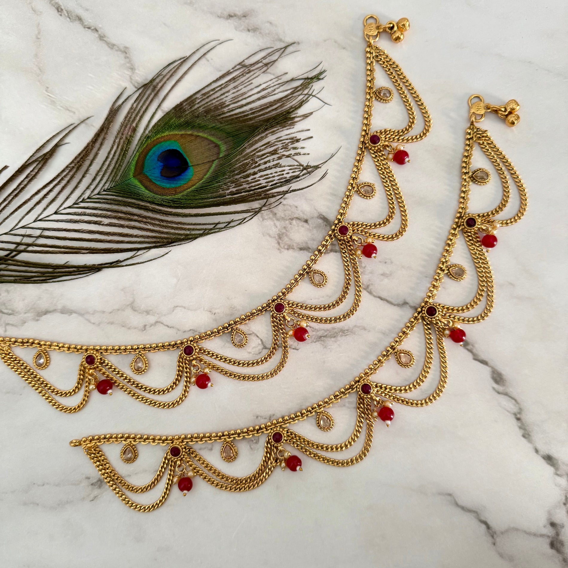 Gold anklet with red stones on a marble surface with a peacock feather.