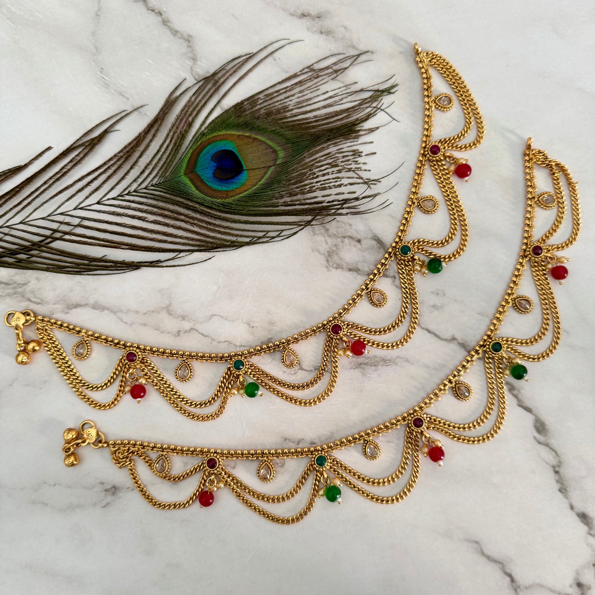 Gold anklet with colorful stones on a marble surface with a peacock feather.