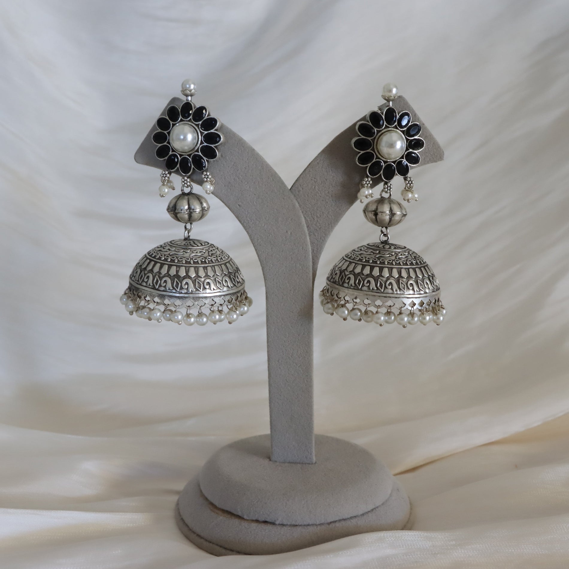 A pair of traditional Indian silver jhumka earrings with black enamel flower details, white faux pearls, and silver beadwork, displayed on a gray velvet jewelry stand against a draped white fabric background.
