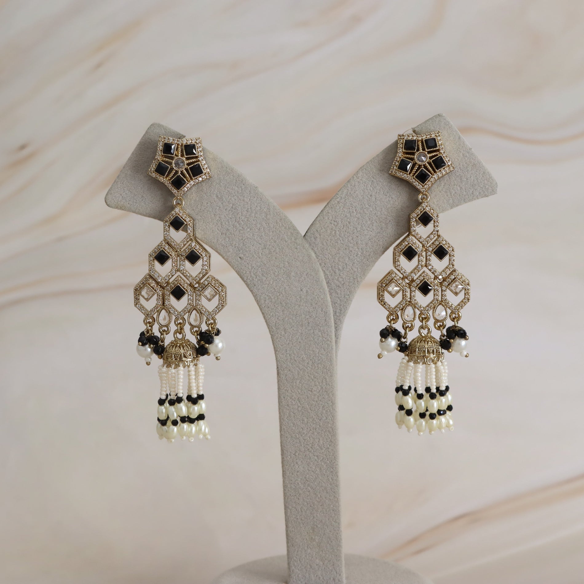 A pair of ornate gold-toned dangle earrings with black and white beadwork, displayed on a light gray jewelry stand against a soft beige marble background.