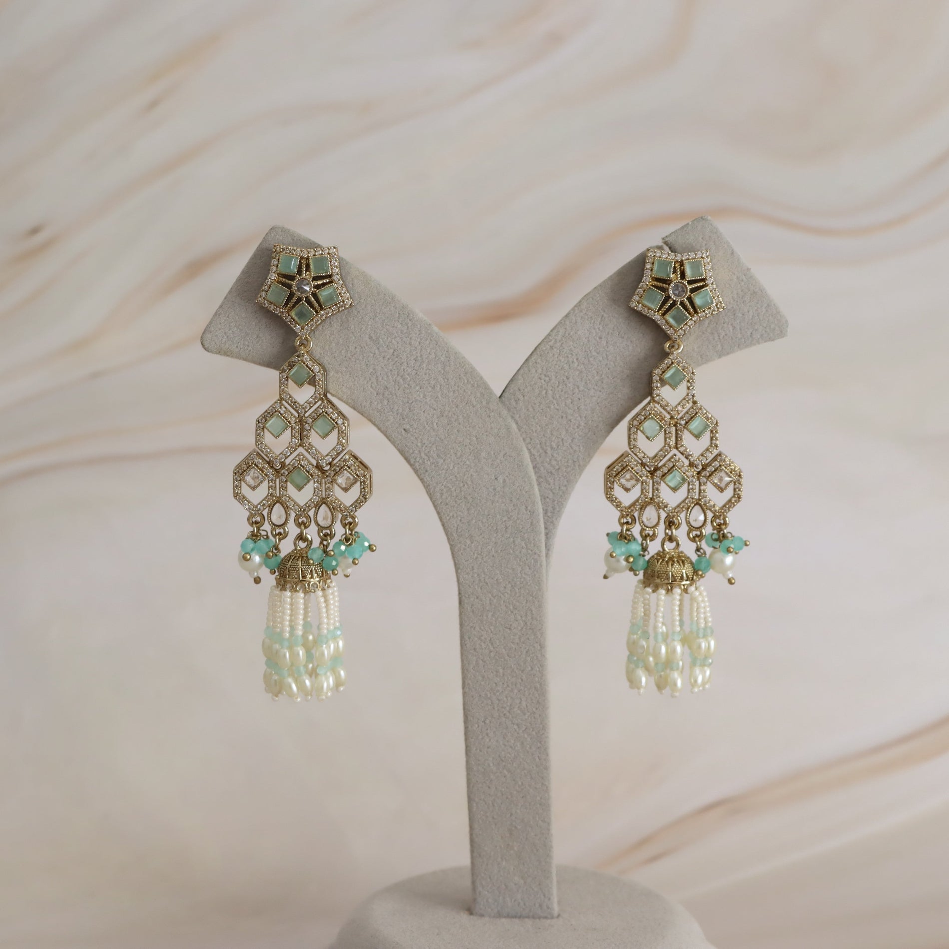 A pair of ornate gold-toned statement earrings with mint green stones and white pearl tassel drops, displayed on a light gray Y-shaped jewelry stand against a soft beige marble background.