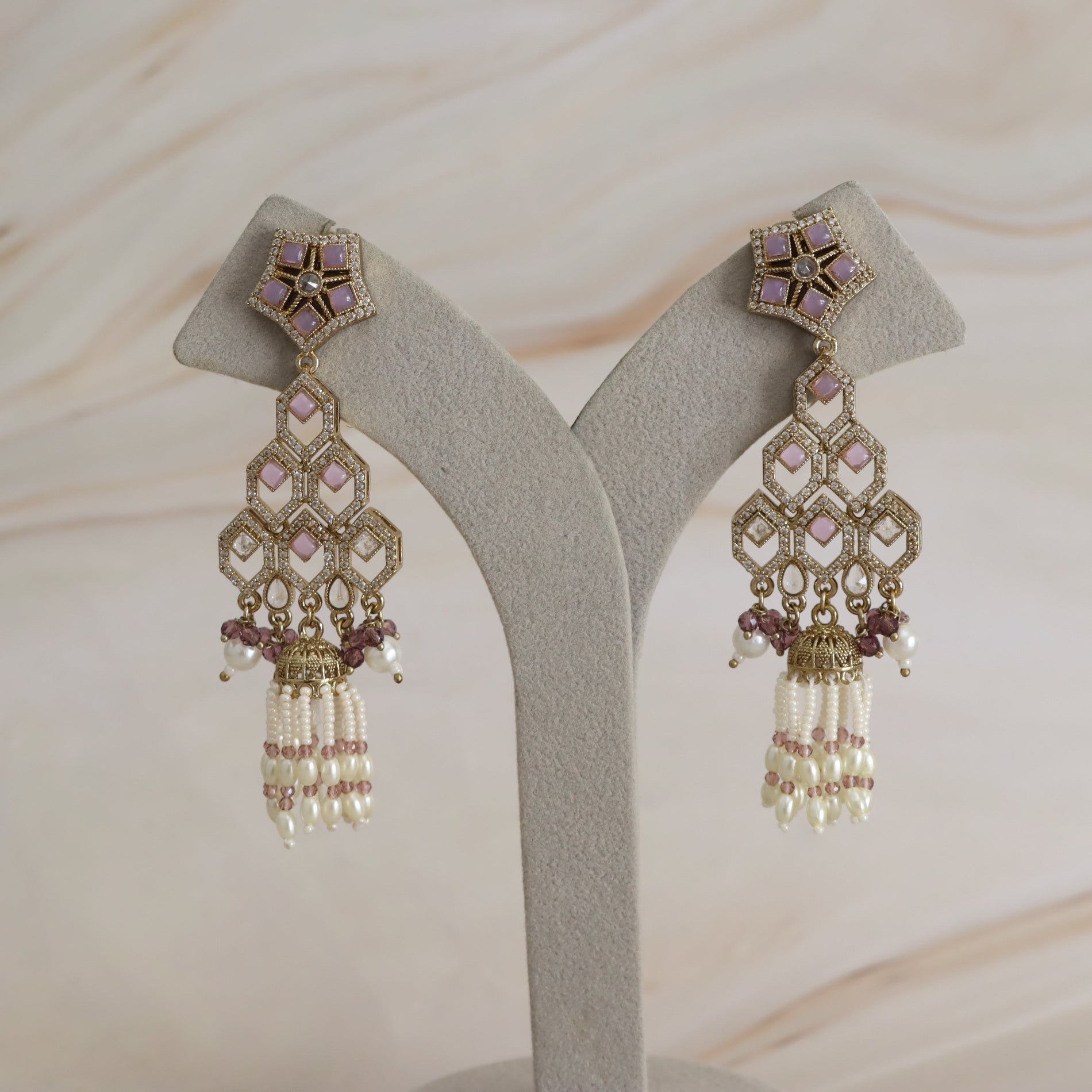 A pair of ornate gold-toned dangle earrings with pink and white stones and pearl-like drops, displayed on a light gray jewelry stand against a soft beige marble background.