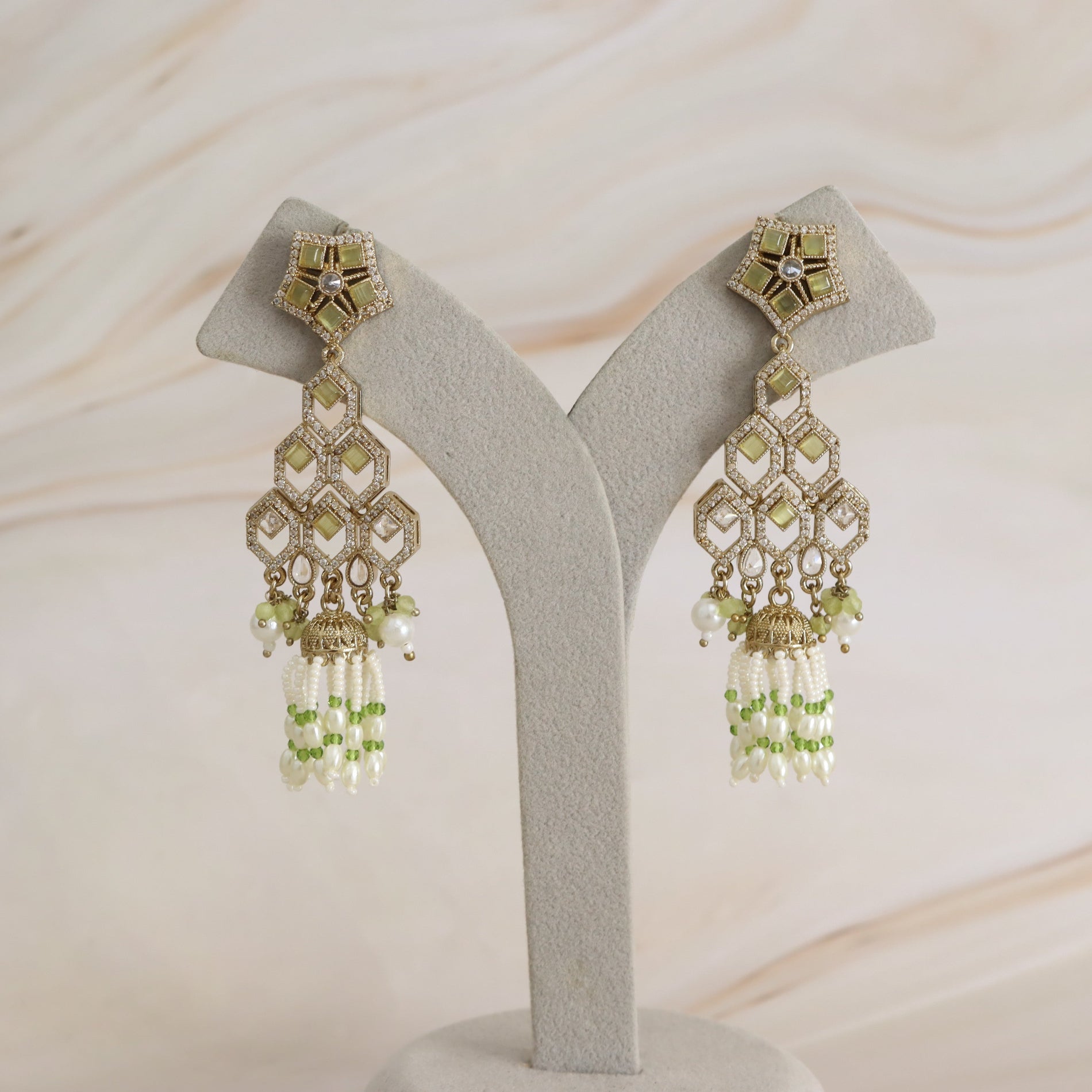 A pair of ornate gold-toned statement earrings with light green stones, white pearl-like beads, and dangling green bead tassels, displayed on a gray jewelry stand against a soft beige marble background.