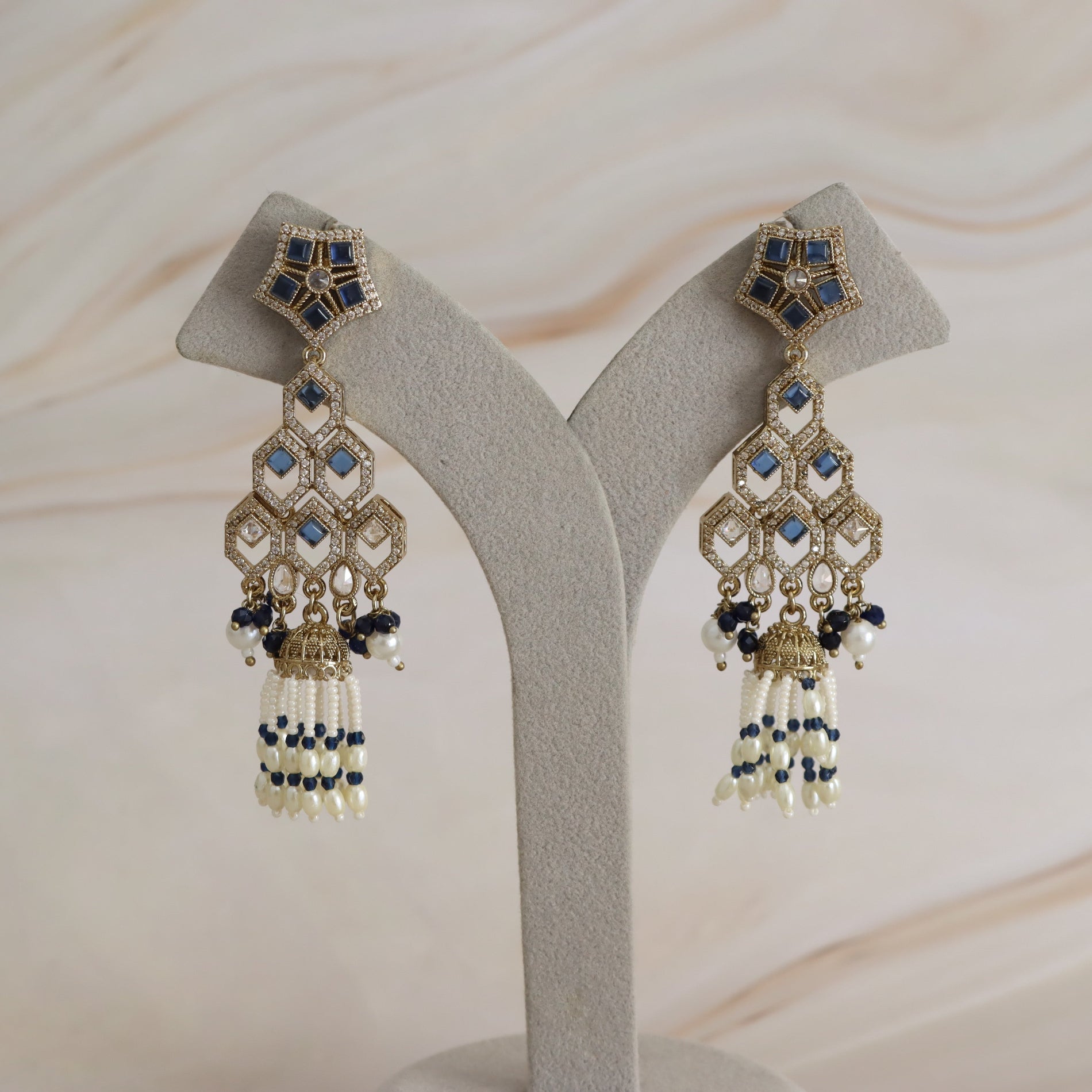 A pair of ornate gold-toned statement earrings with blue and white beadwork, displayed on a light gray jewelry stand against a soft beige marble background.