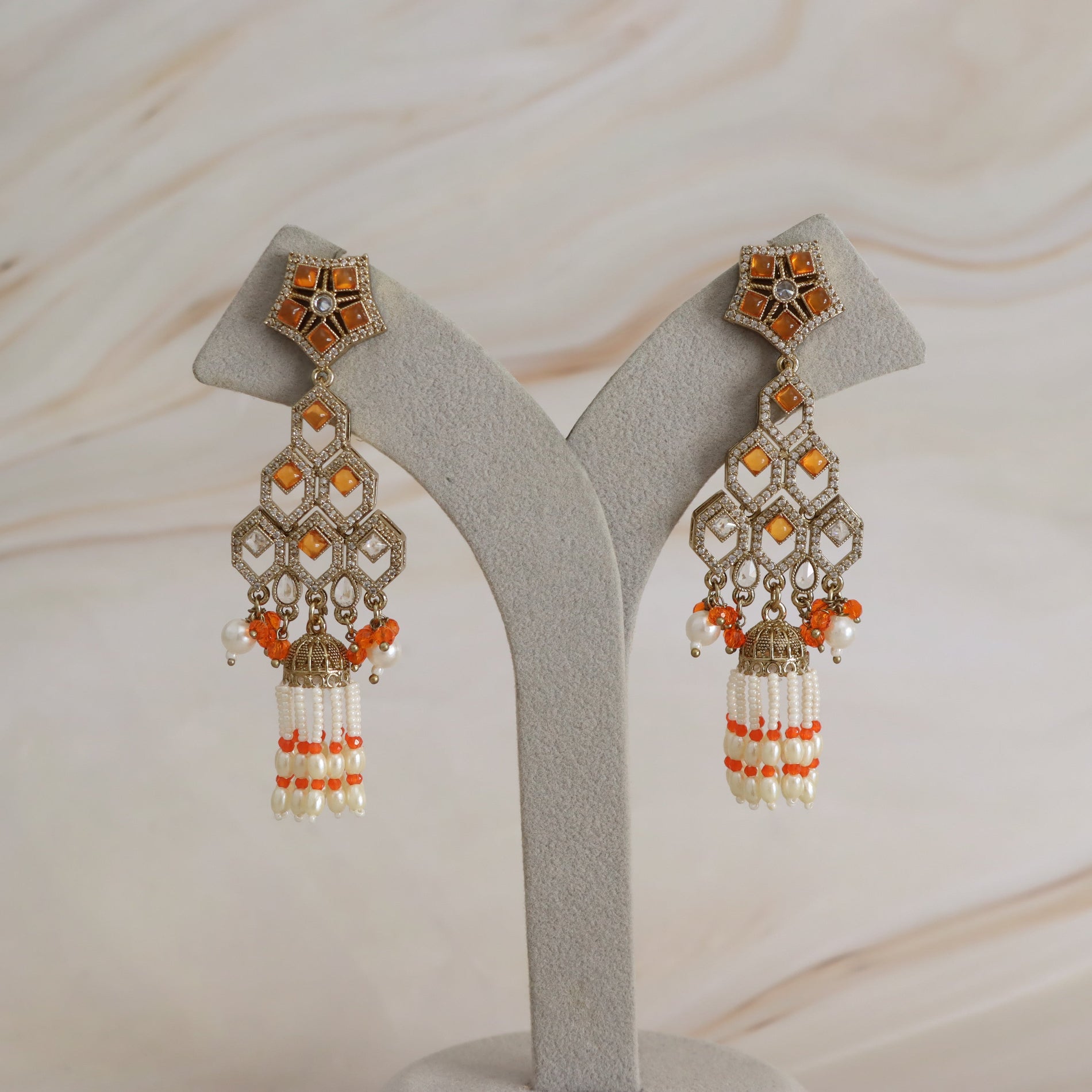 A pair of ornate gold-toned dangle earrings with orange and white stones and bead tassels, displayed on a light gray jewelry stand against a soft beige marble background.