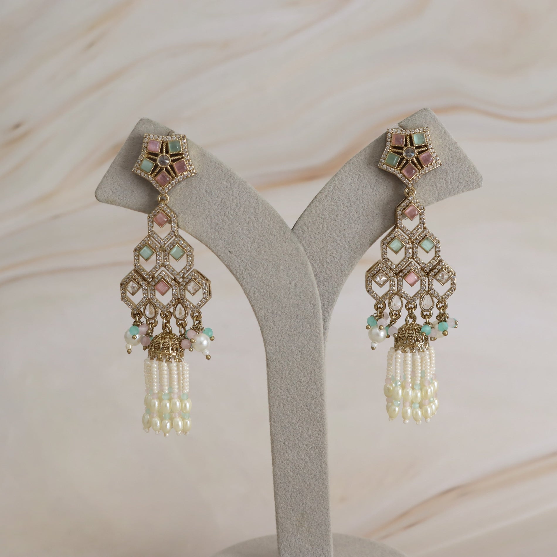 A pair of ornate gold-toned dangle earrings with pastel pink, mint green, and white stones, displayed on a gray jewelry stand against a light beige marble background.