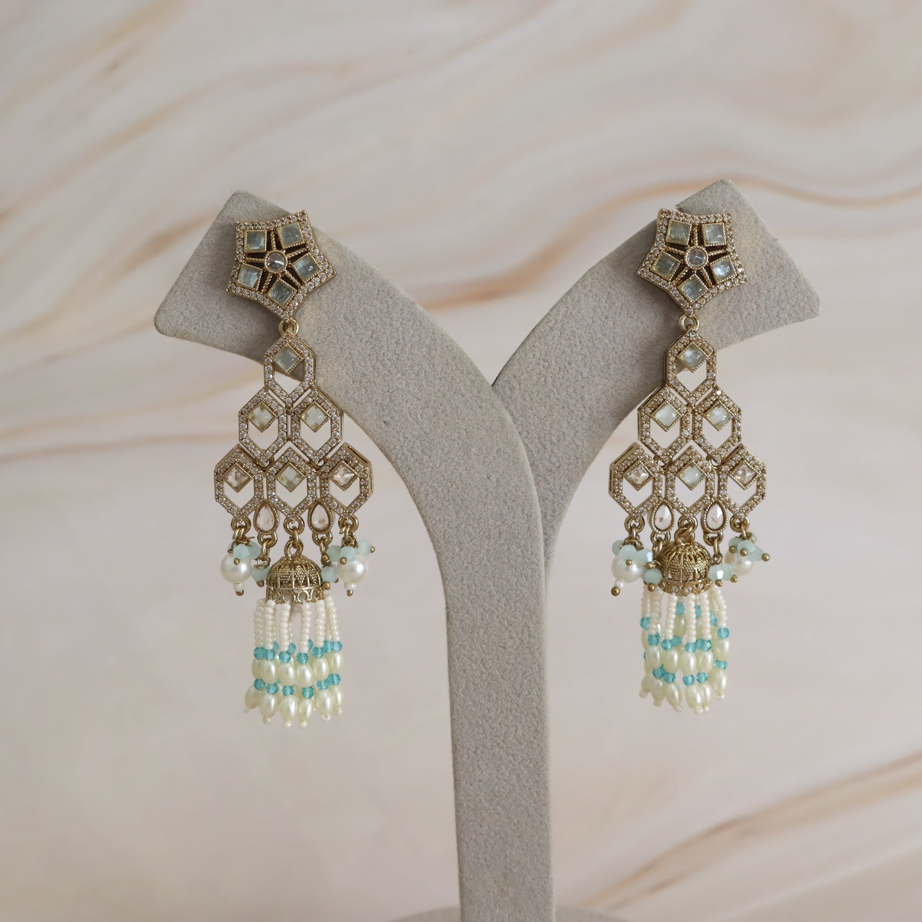 A pair of ornate gold-toned statement earrings with white pearl-like beads and light blue accents, displayed on a gray velvet jewelry stand against a light beige marble background.